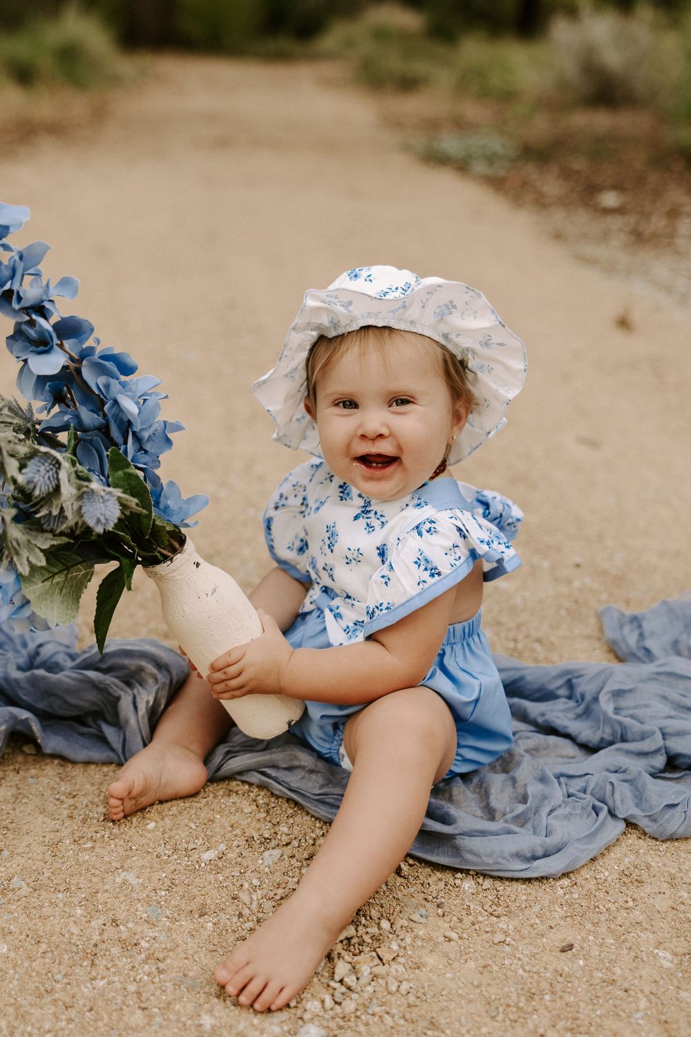 A small child in a blue and white outfit sits on a blanket outdoors holding a bottle next to blue flowers.