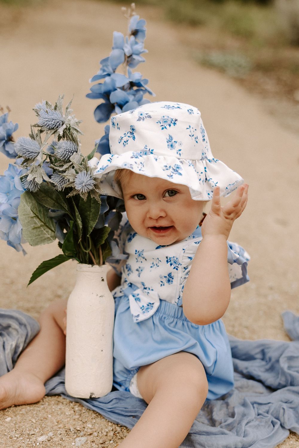 A baby in a floral bonnet lies on a blanket with blue flowers and white vase decorations.