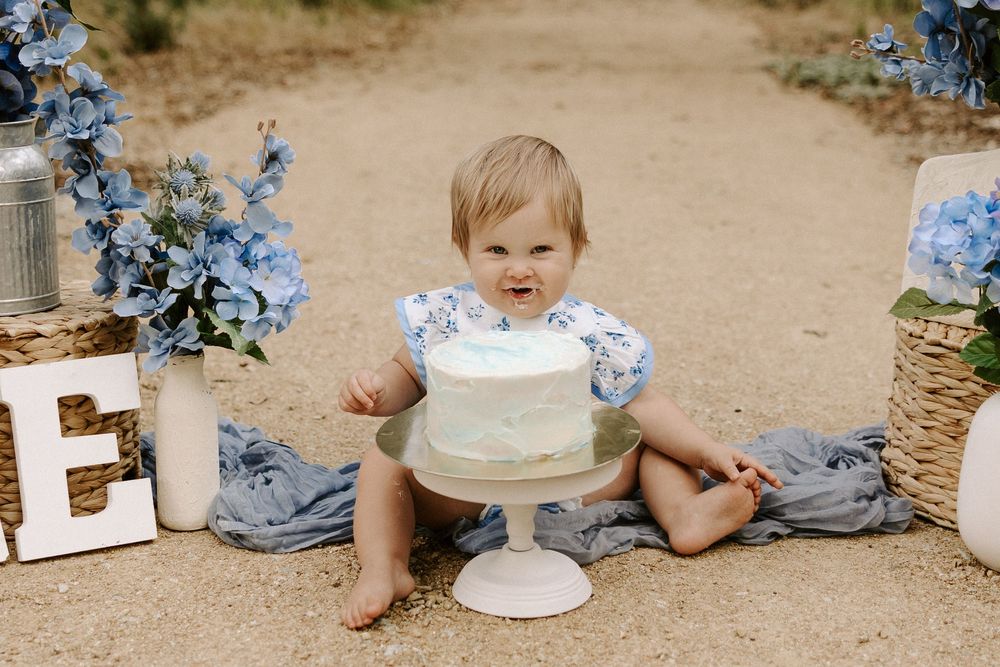 A baby in a blue outfit sits on the ground near a white cake stand and delicate blue hydrangea flowers.