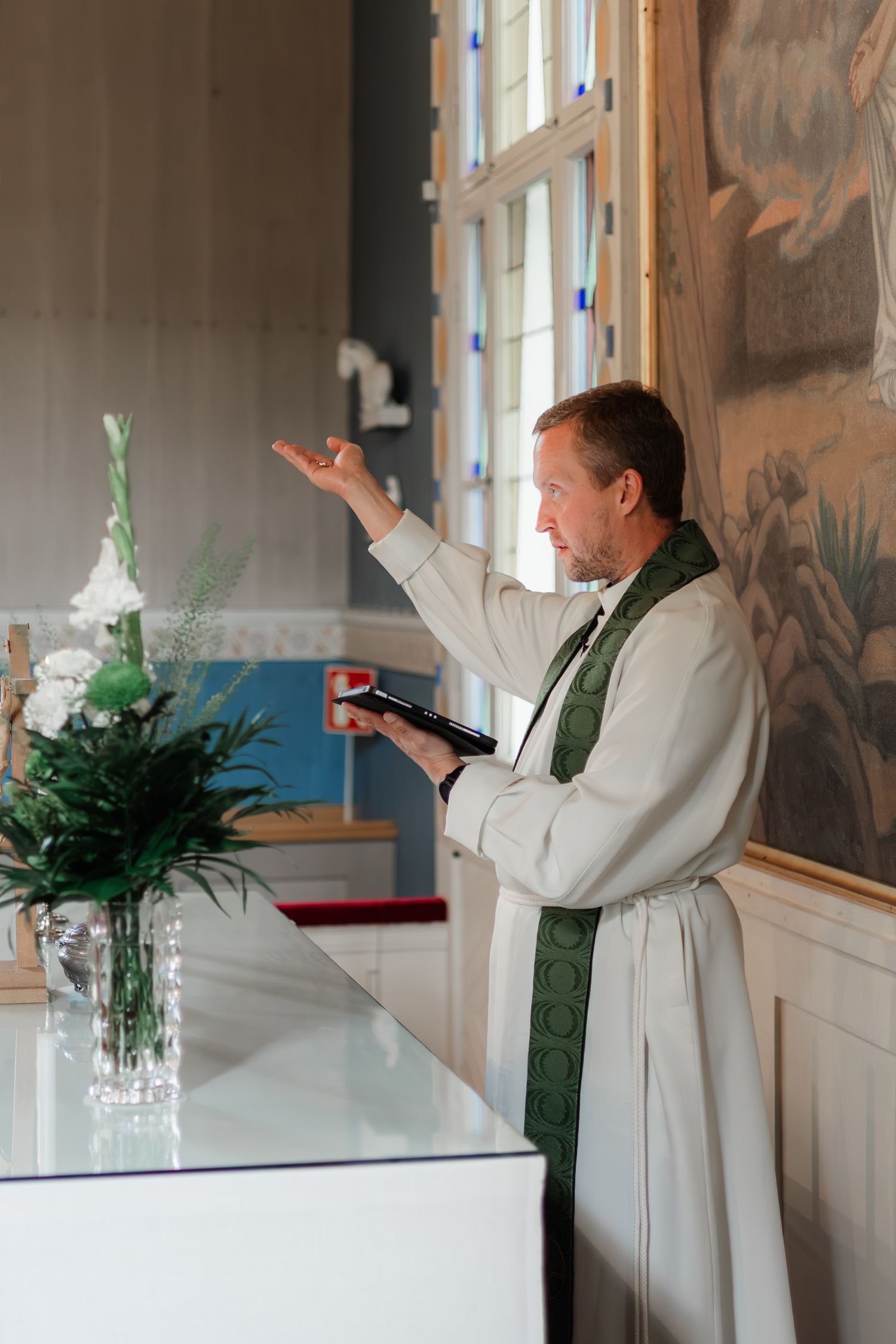 A clergy member in white robes and green stole conducts a religious service at an altar with floral decorations.