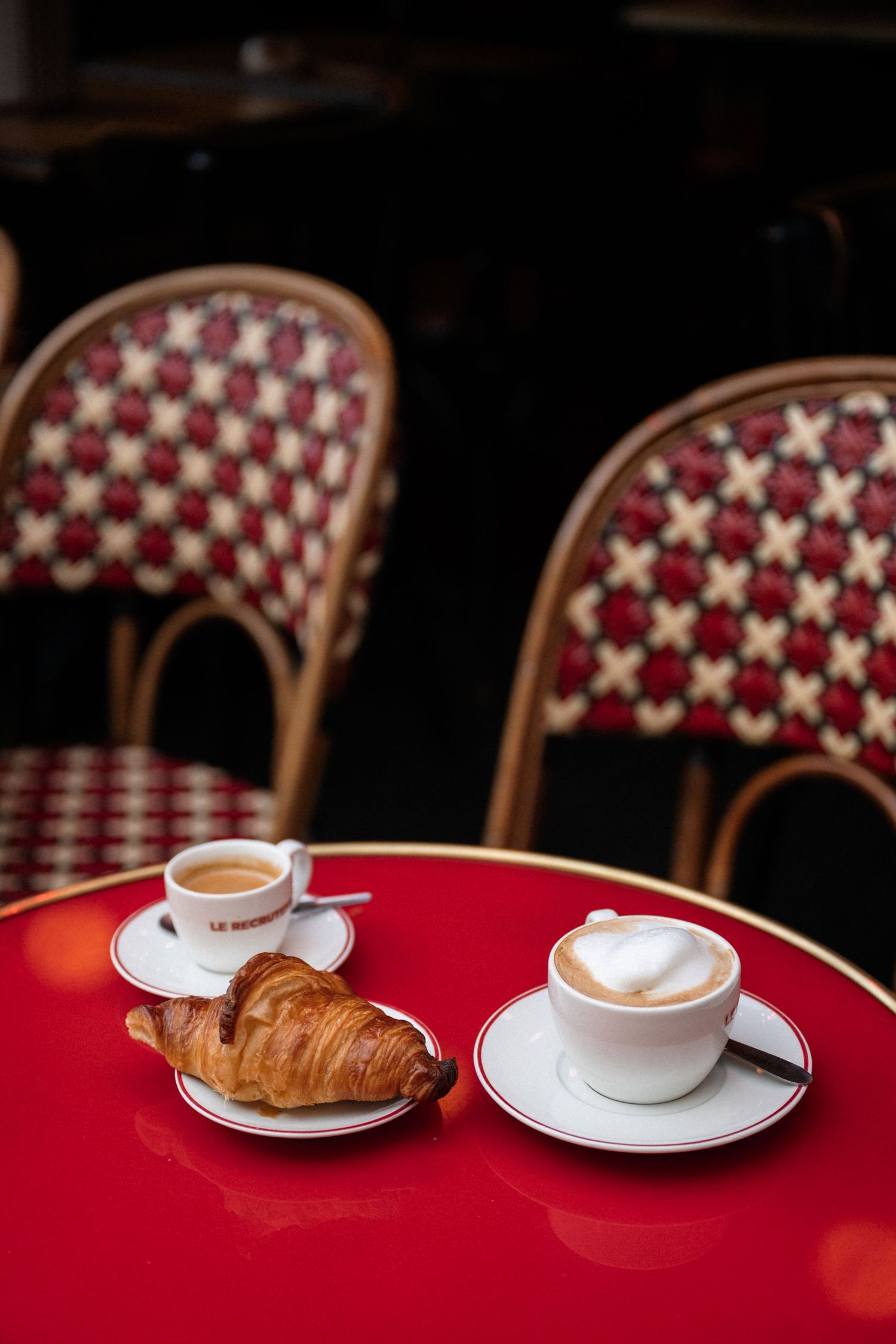 A coffee cup and croissant sit on a round red table with decorative woven chairs visible in the background.