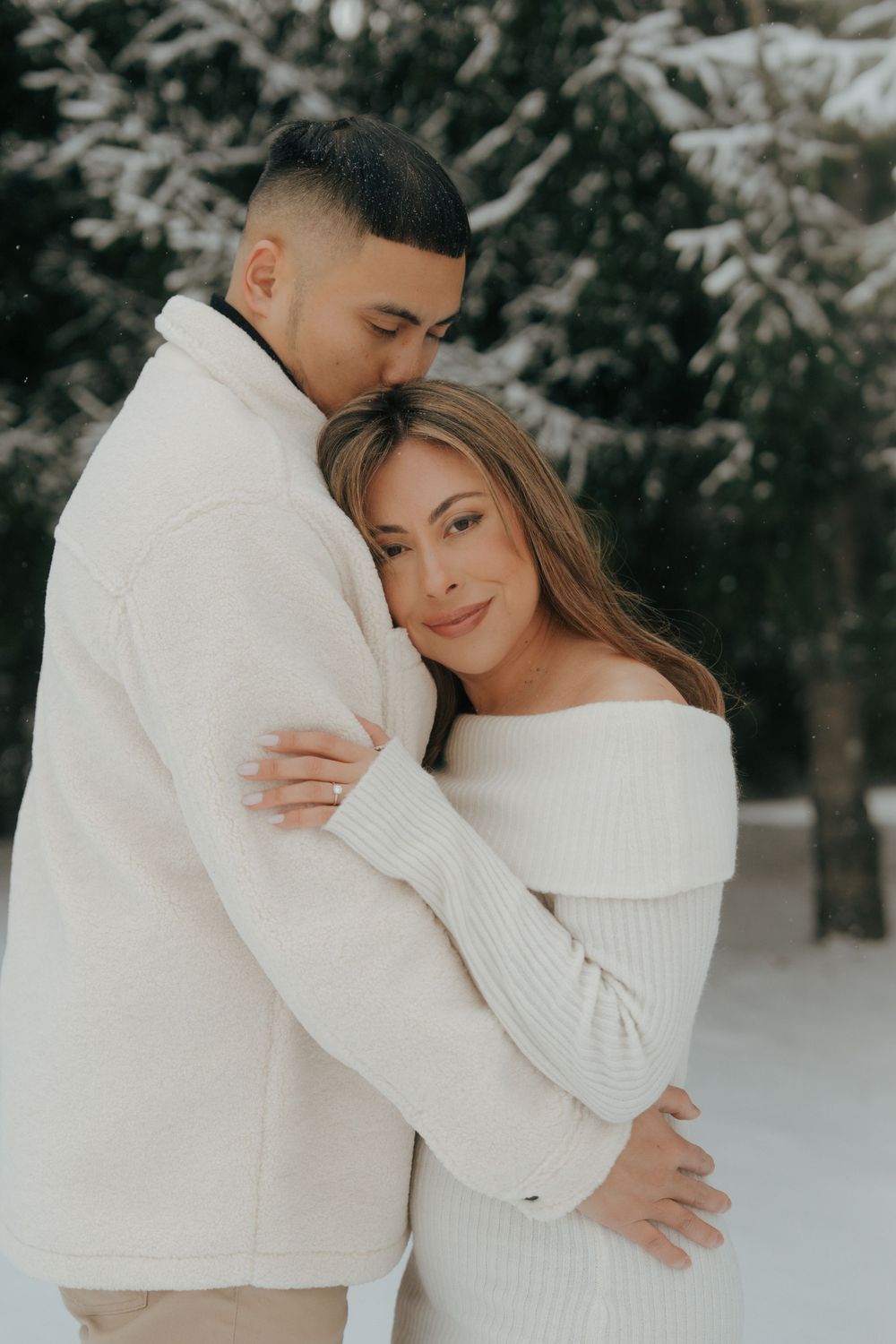 A romantic winter engagement photo series showing a couple in white sweaters embracing in the snow.