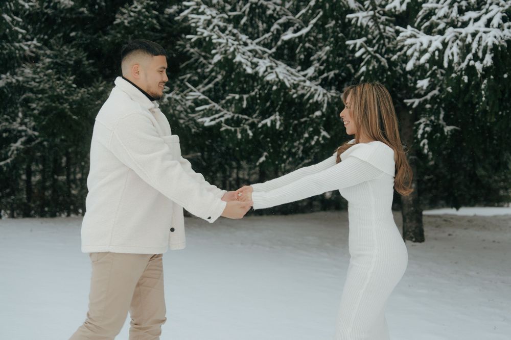 A couple in matching white outfits holds hands while standing in a snowy winter forest setting.