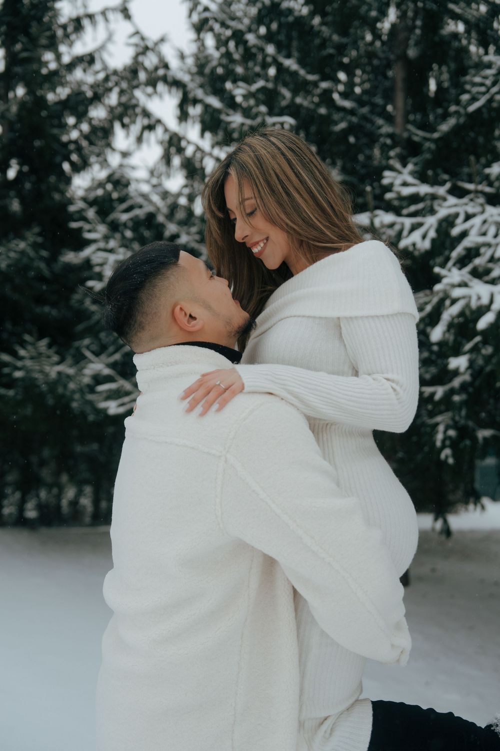 A couple wearing white sweaters embrace lovingly in a snowy winter forest setting.