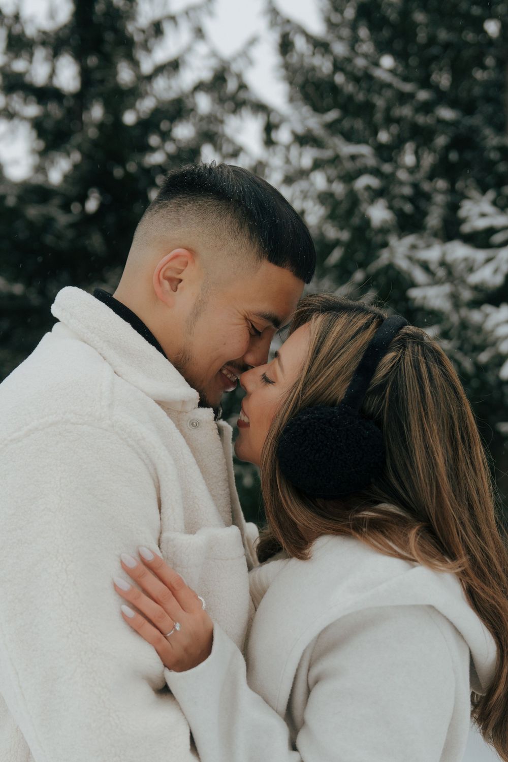 A romantic winter scene shows a couple in cozy white sweaters sharing an intimate moment among snow-covered evergreens.