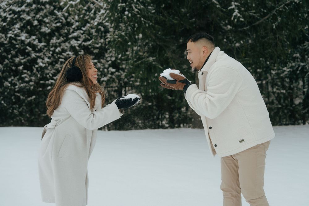Couple walking together through snowy winter forest holding hands in matching white and beige outfits.