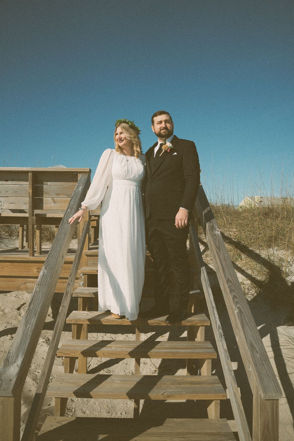 A couple in wedding attire sits together on wooden beach boardwalk steps against a bright blue sky. Wedding Photographer in Carolina Beach