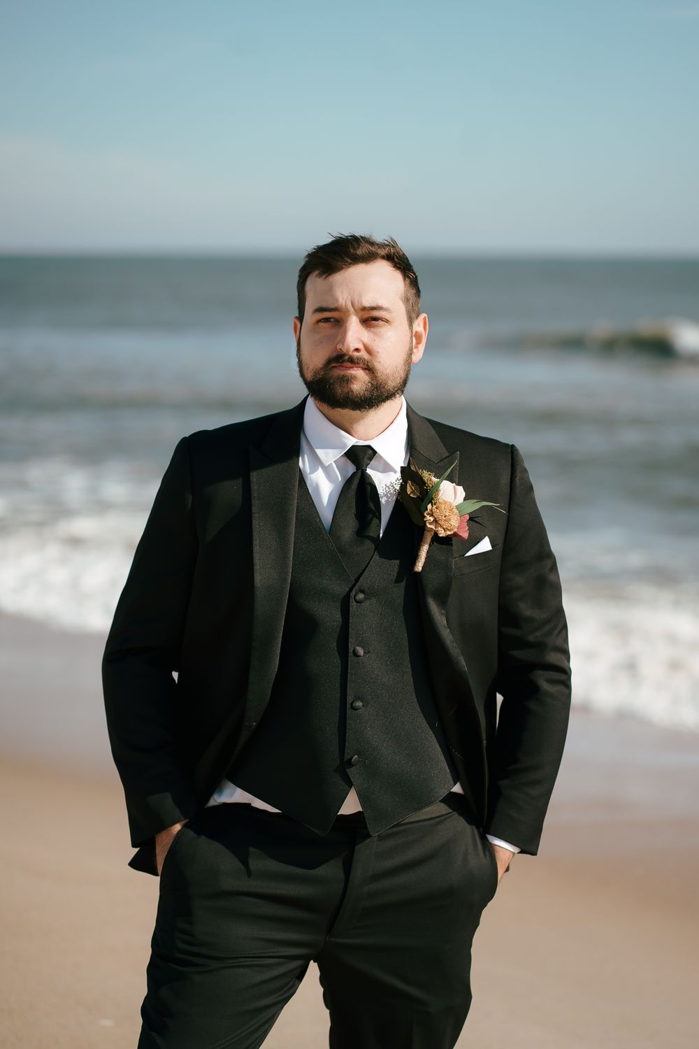 A groom in a classic black  suit with boutonniere stands confidently on a beach with ocean waves behind him. Wilmington, NC Wedding