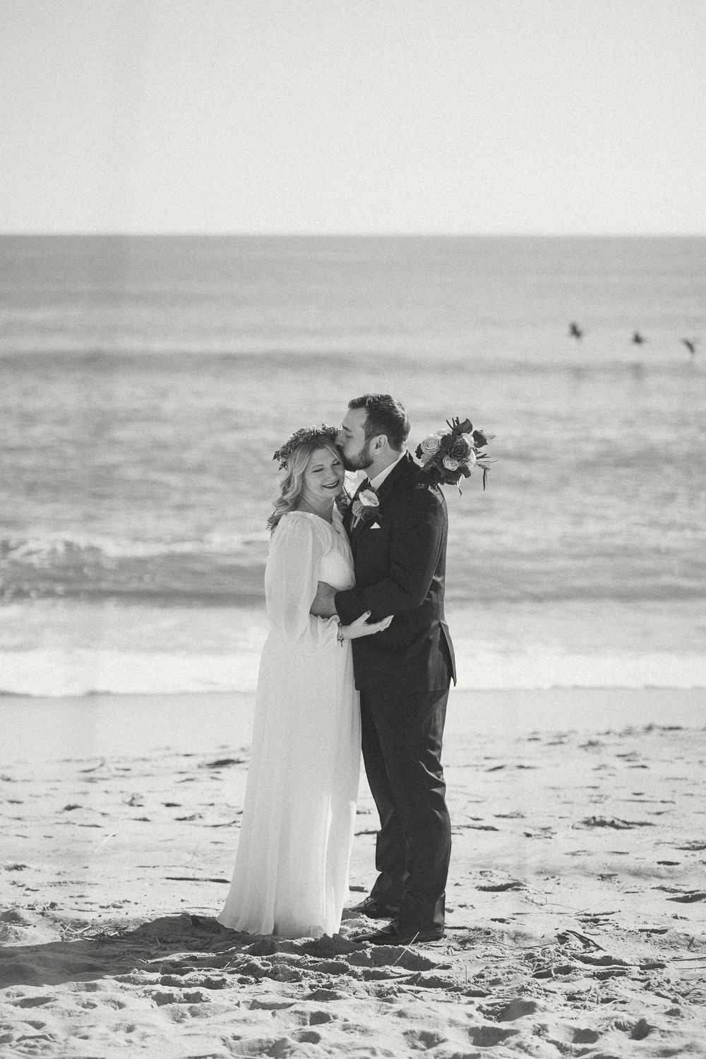 A romantic black and white beach wedding photo shows a couple embracing on the sand with ocean waves in the background. Carolina Beach, NC