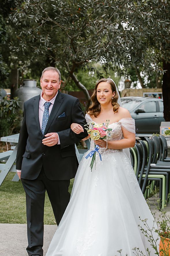 A bride in a white off-shoulder gown walks down an outdoor path holding a pink bouquet, accompanied by a man in a suit.