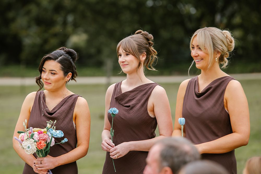 A row of bridesmaids in brown dresses stand together outdoors holding small bouquets during a wedding ceremony.