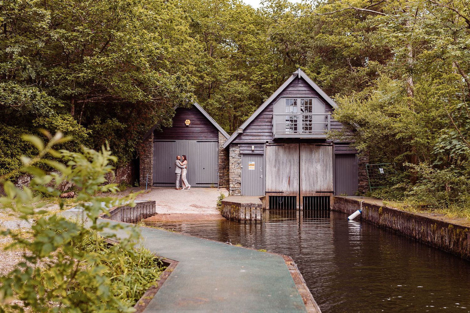 Historic wooden boathouses line a tranquil river surrounded by lush green trees and foliage in summer.