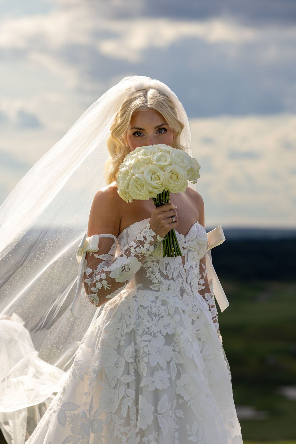 Blonde bride in lace off-shoulder gown holds white rose bouquet with veil flowing in wind against cloudy sky.