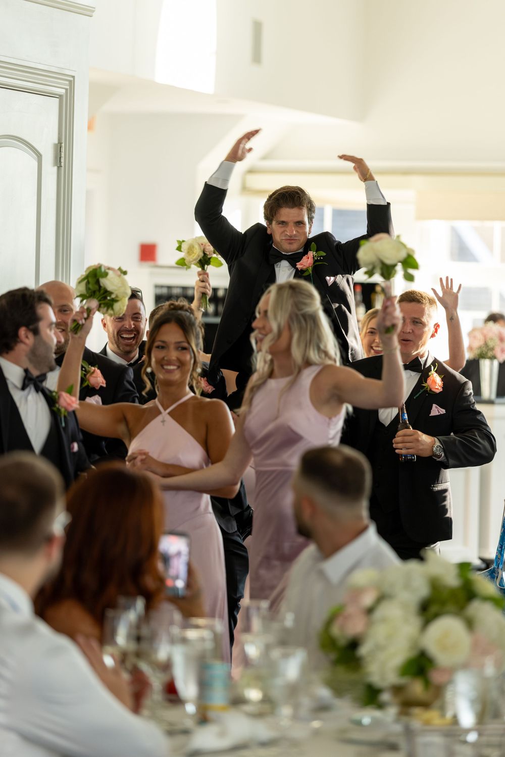Joyful wedding reception celebration with guests dancing and cheering as groom raises arms triumphantly in elegant venue.