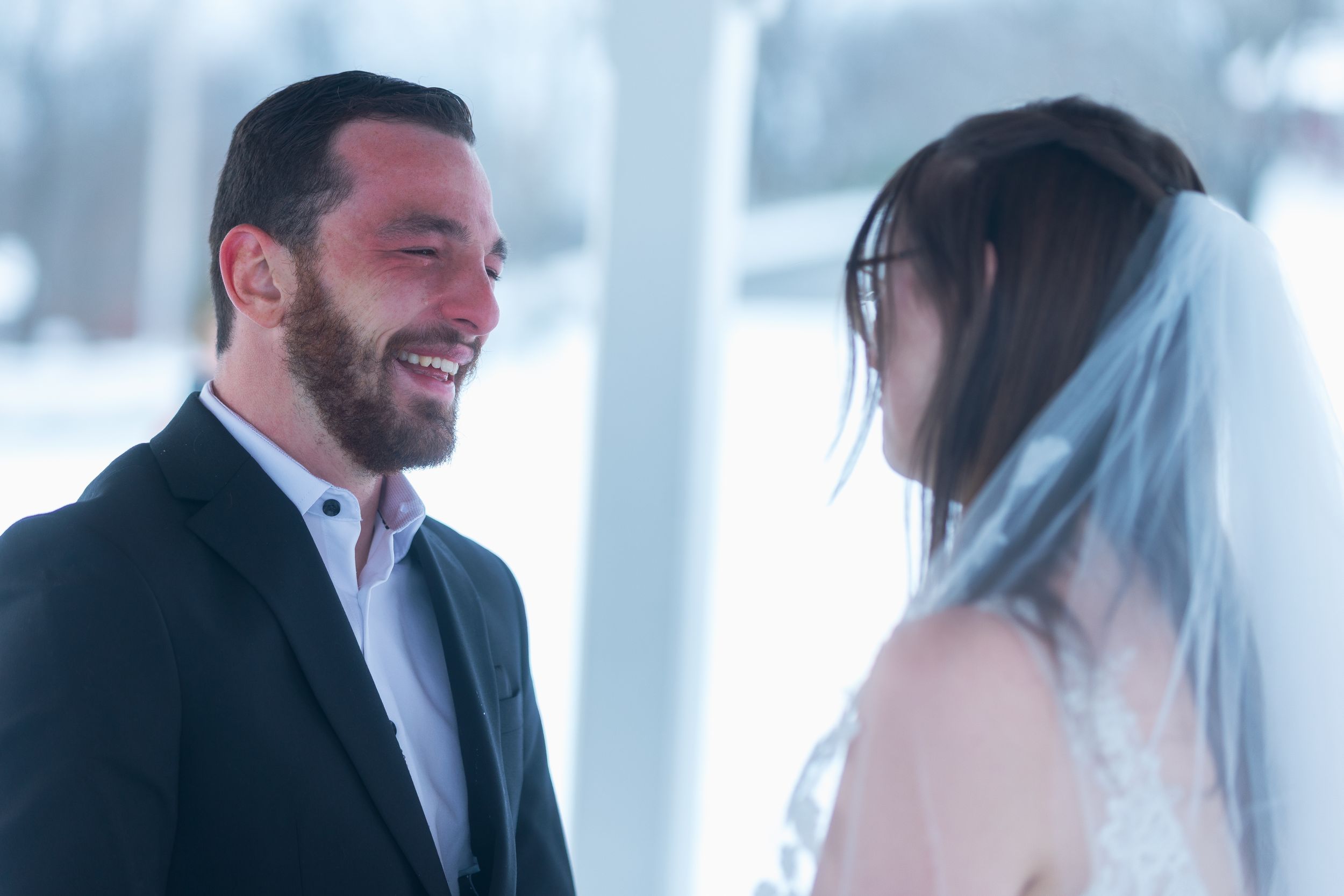 A couple in formal attire stands together in a bright white interior setting during a wedding celebration.