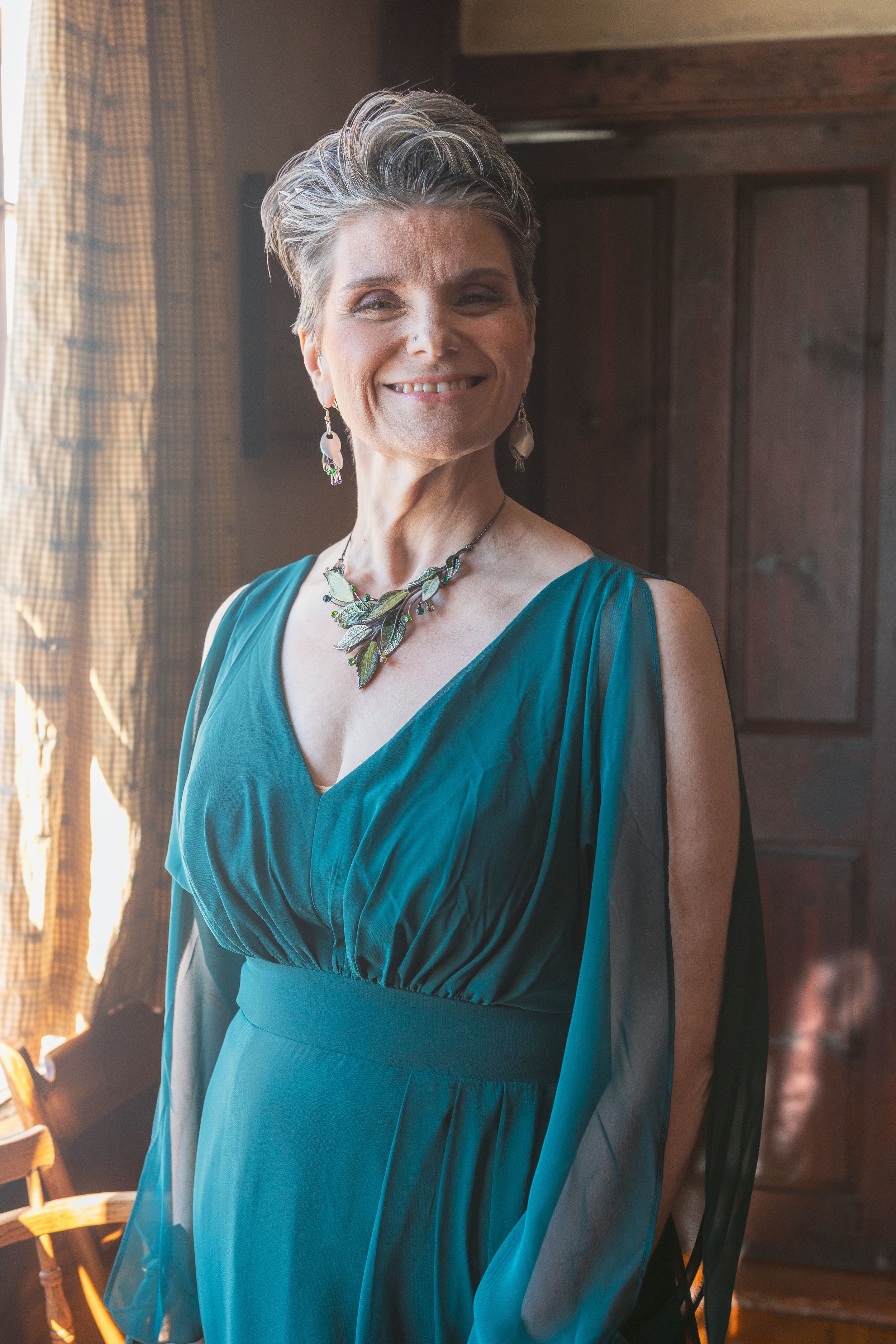 Elegant woman in a teal formal dress with statement necklace stands in warm sunlit room near window.