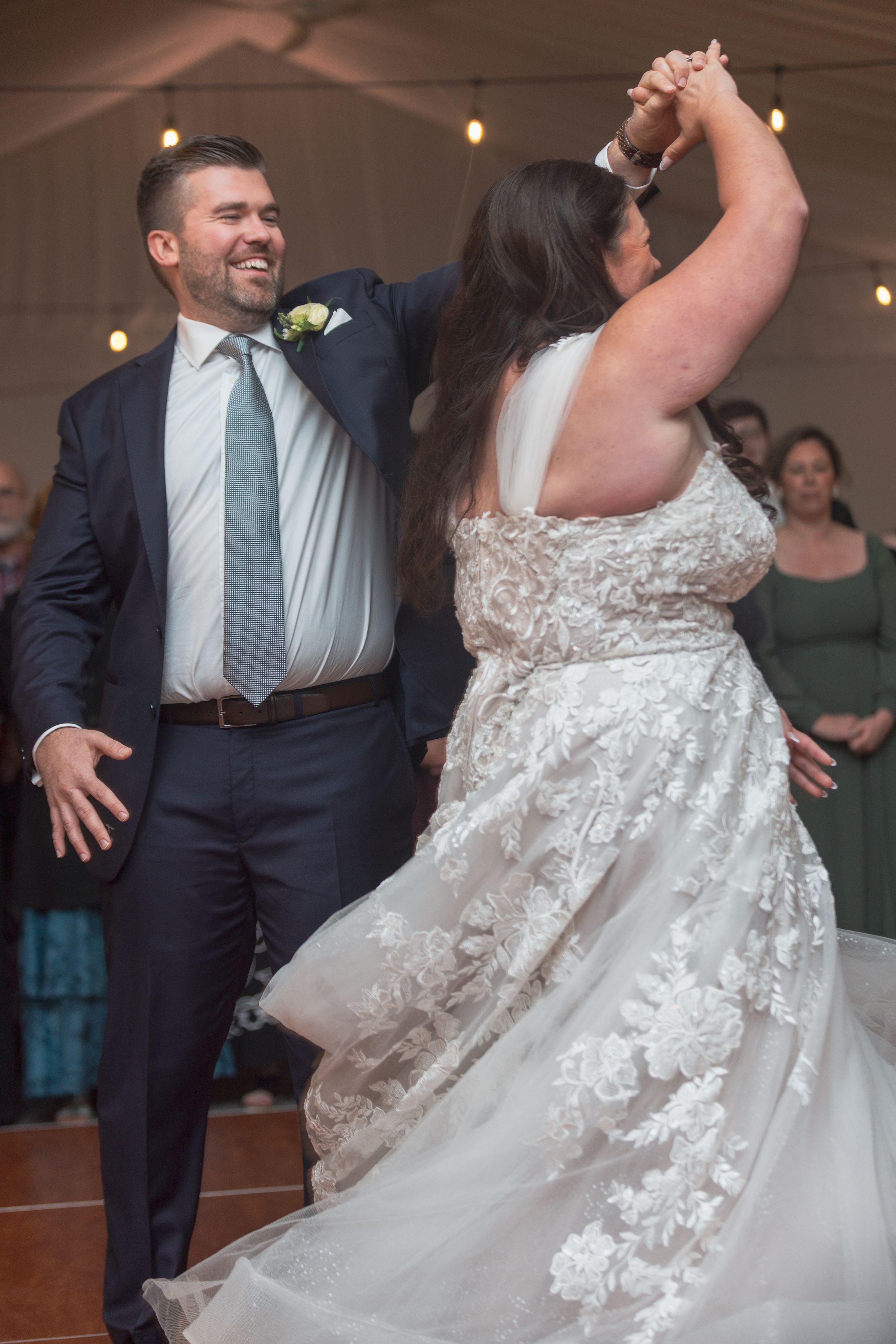 A couple dances together at their wedding reception, with the bride wearing a flowing white lace gown and the groom in a suit.