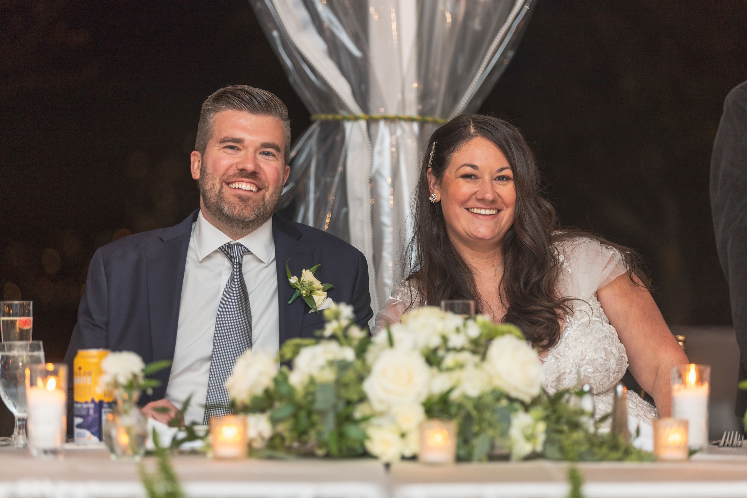 Wedding party laughs together at reception table decorated with white floral centerpieces and glowing candles.