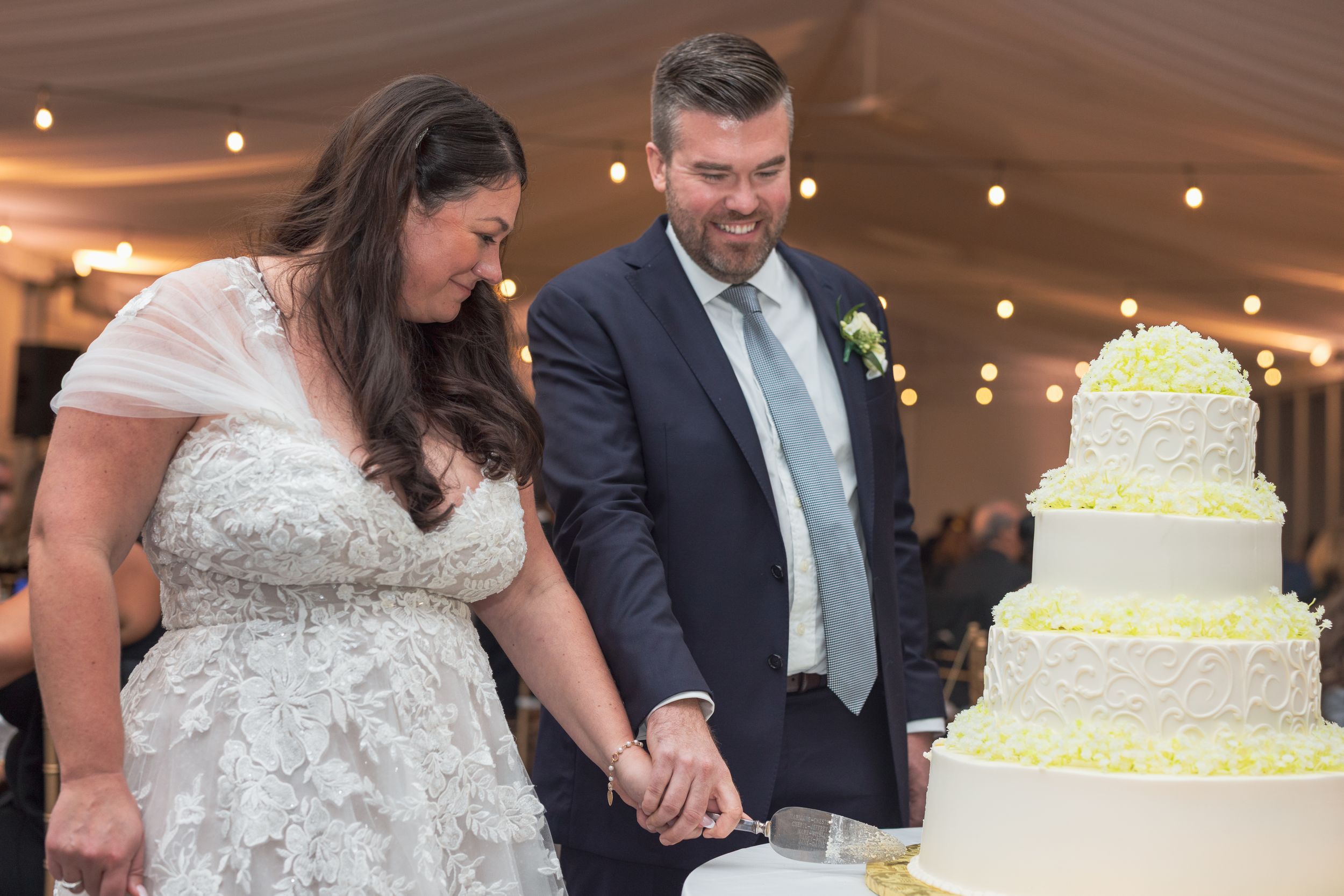 A wedding couple cuts their elegant three-tiered white cake decorated with delicate floral details at their reception.