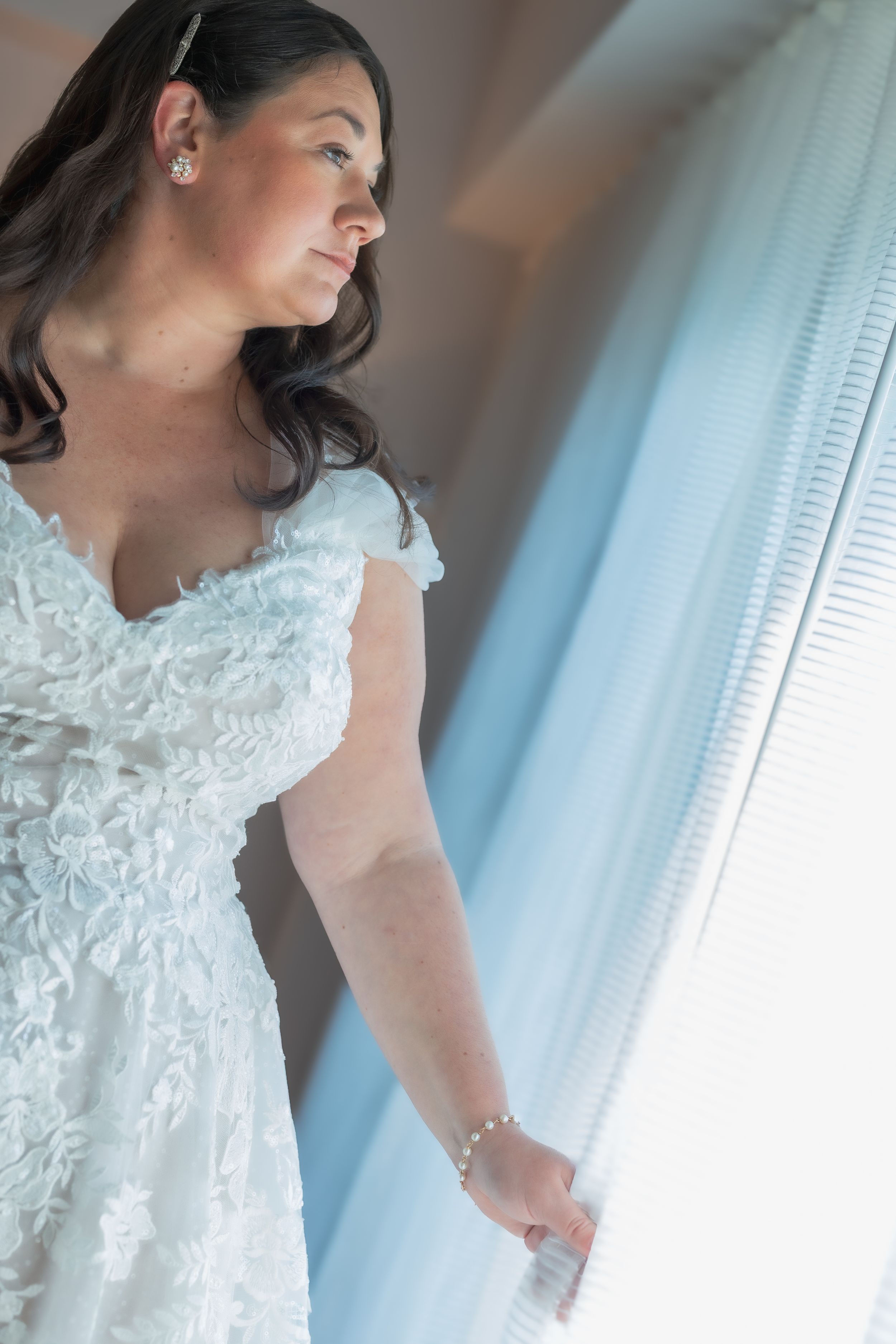 A person in a white lace wedding dress stands by a window with soft blue curtains in natural light.