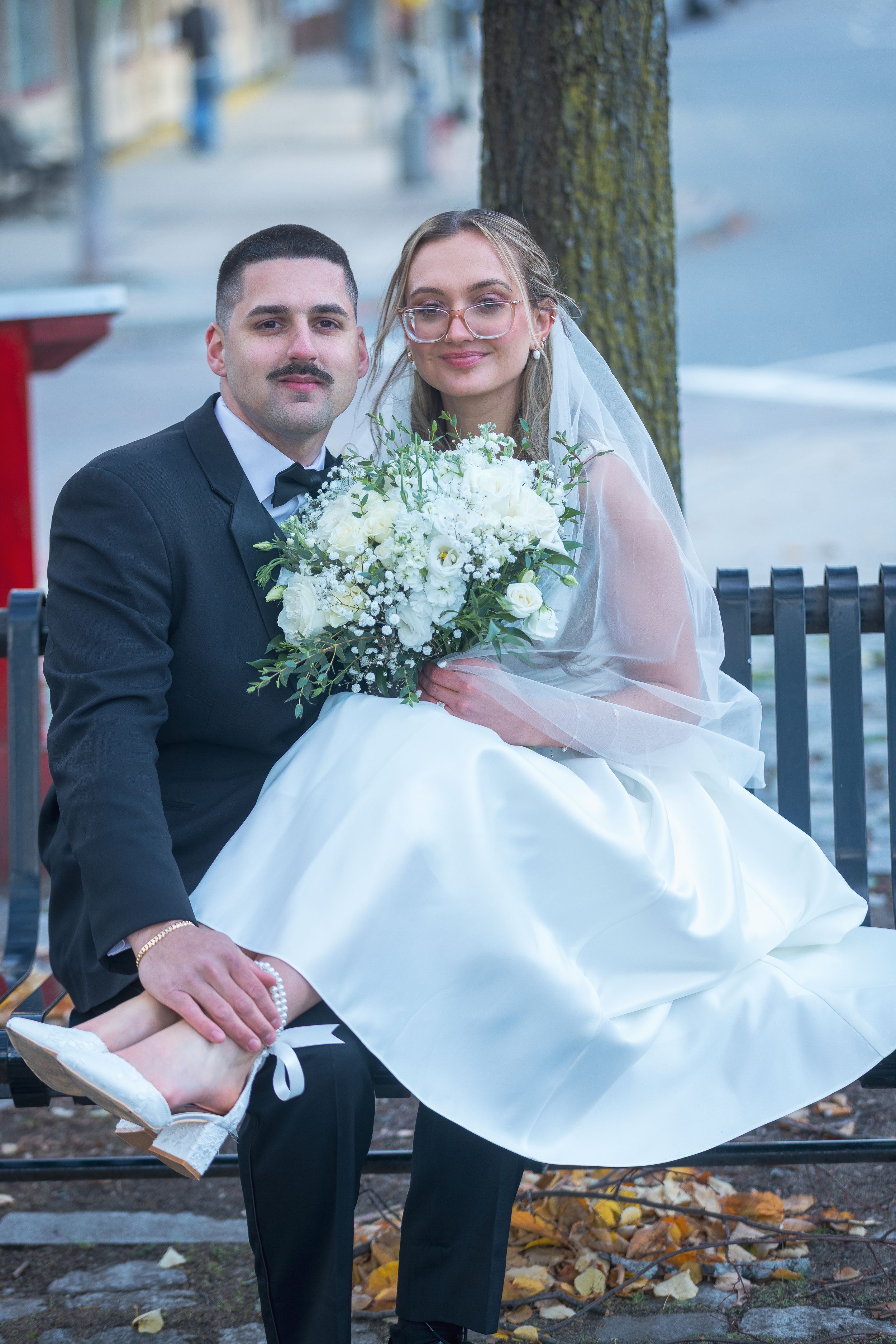 A wedding couple sits on a park bench surrounded by autumn leaves while holding a white floral bouquet.
