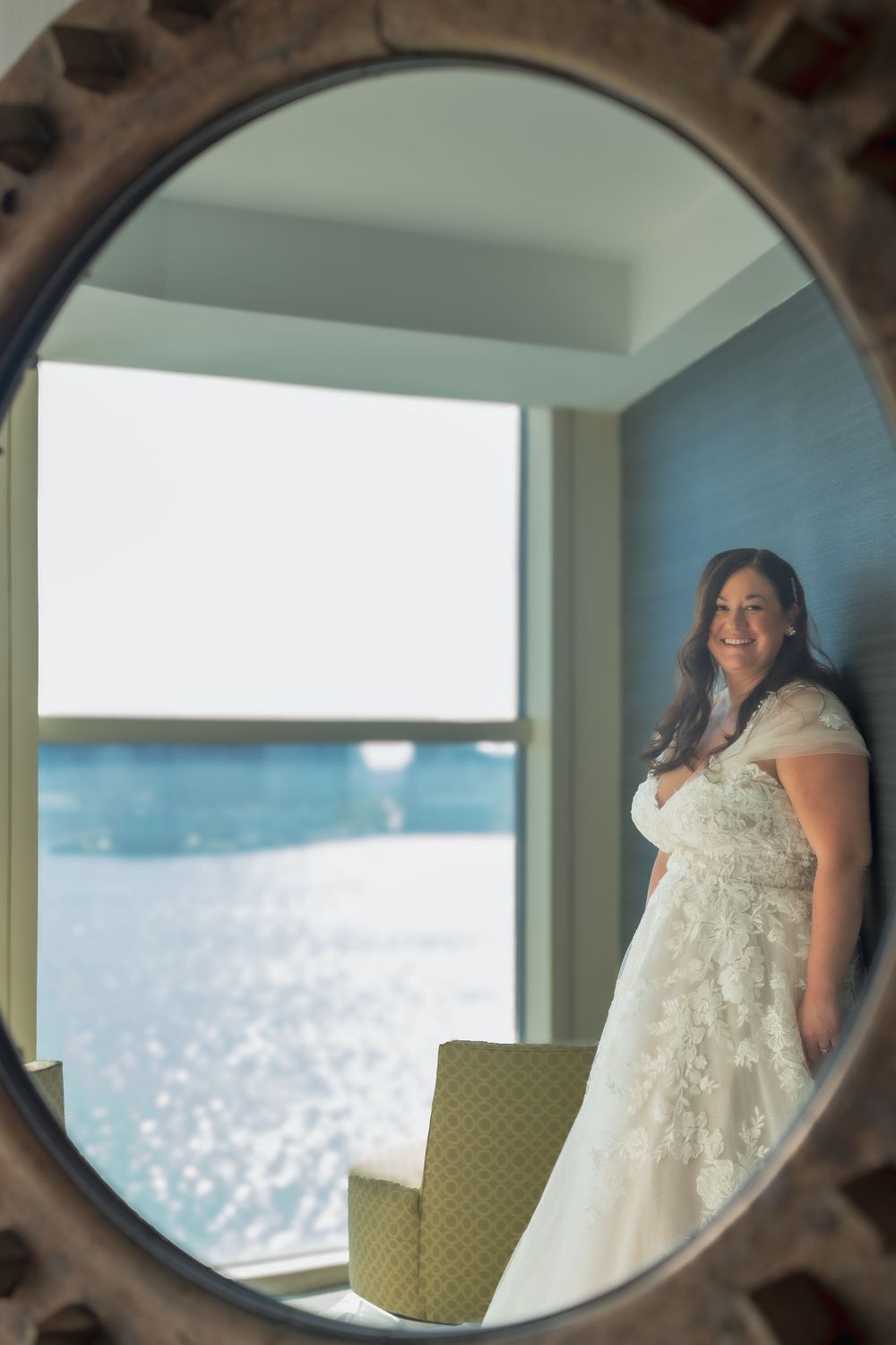 A bride in a white lace wedding dress stands by a window overlooking sparkling ocean waters, viewed through an ornate mirror.