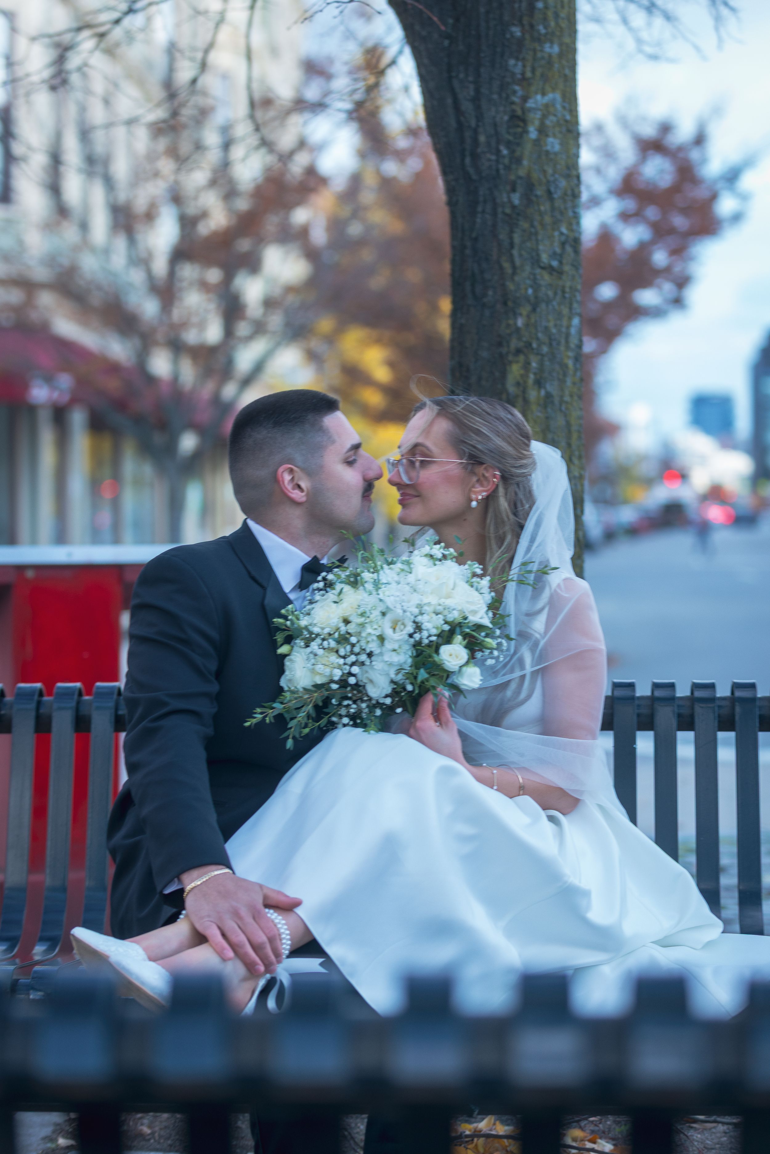 A romantic wedding couple shares tender moments on a park bench with white flowers and autumn scenery in the background.