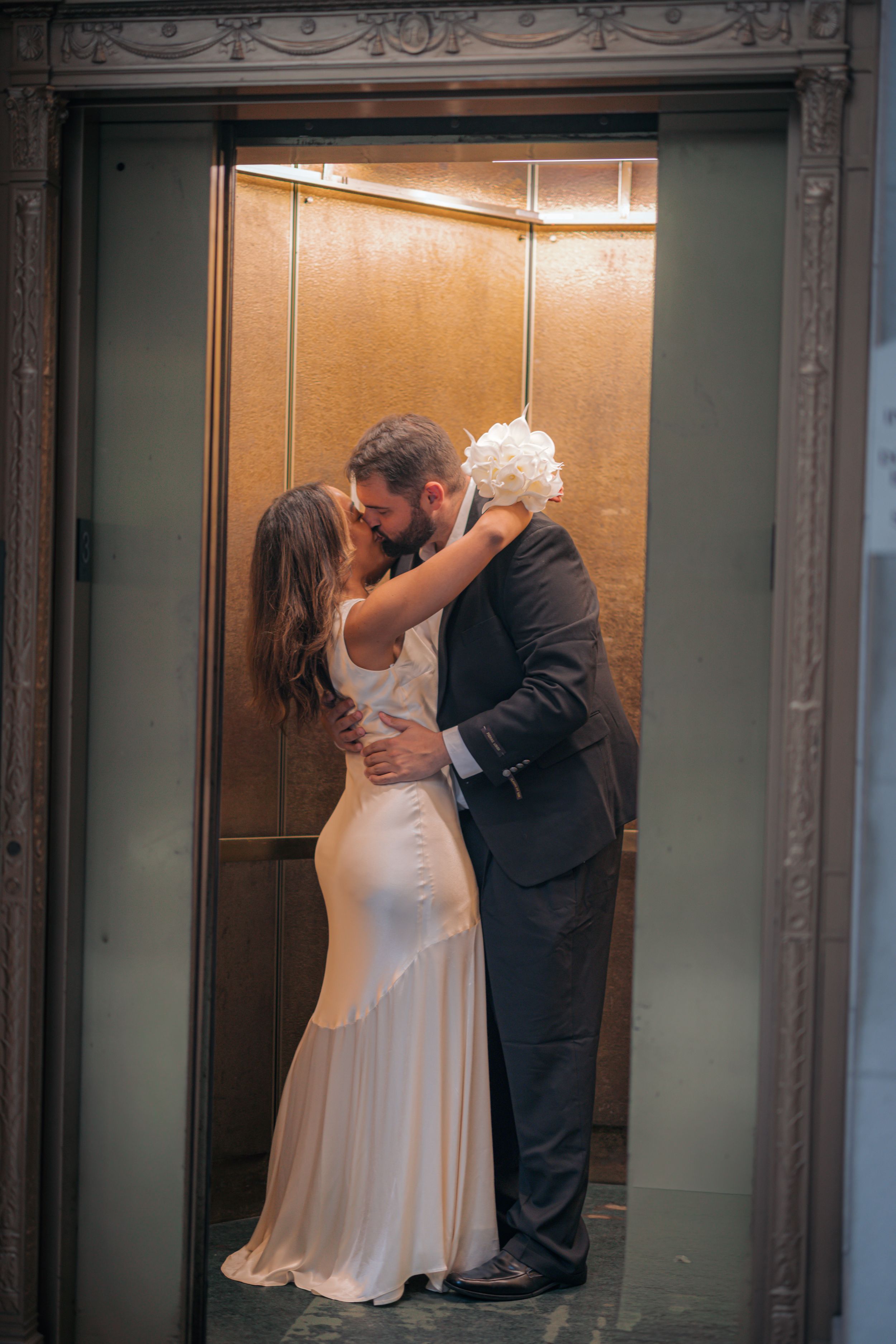A couple shares a romantic kiss in an elevator during their wedding, with the bride in white and groom in black attire.