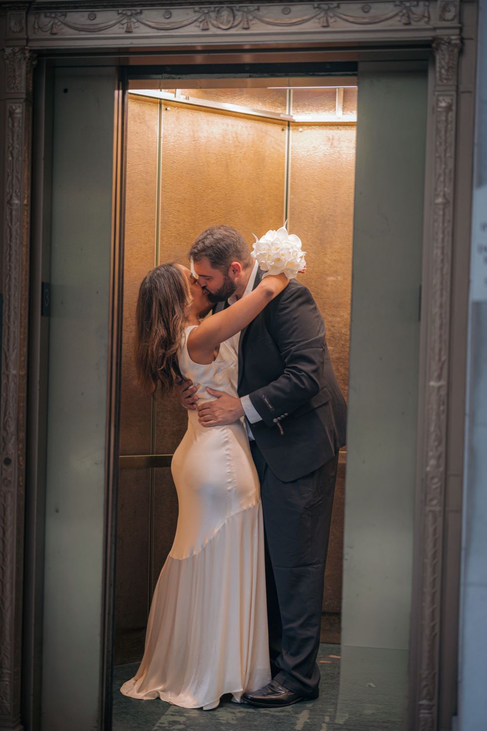 A couple shares a romantic kiss in an elevator during their wedding, with the bride in white and groom in black attire.