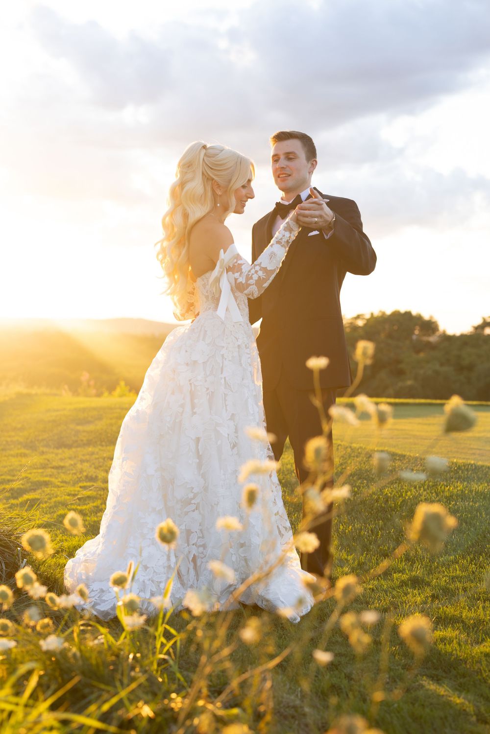 Bride and groom dancing at golden sunset on green meadow, bride in elegant lace gown with flowing train.