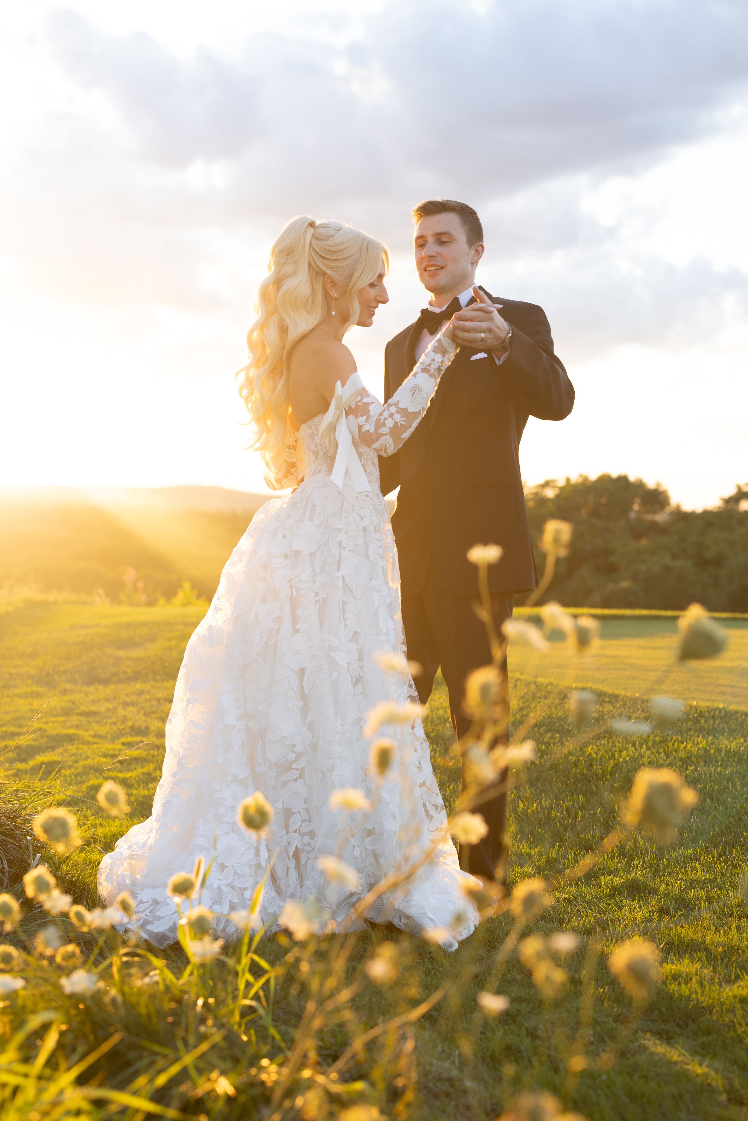 Bride and groom dancing at golden sunset on green meadow, bride in elegant lace gown with flowing train.