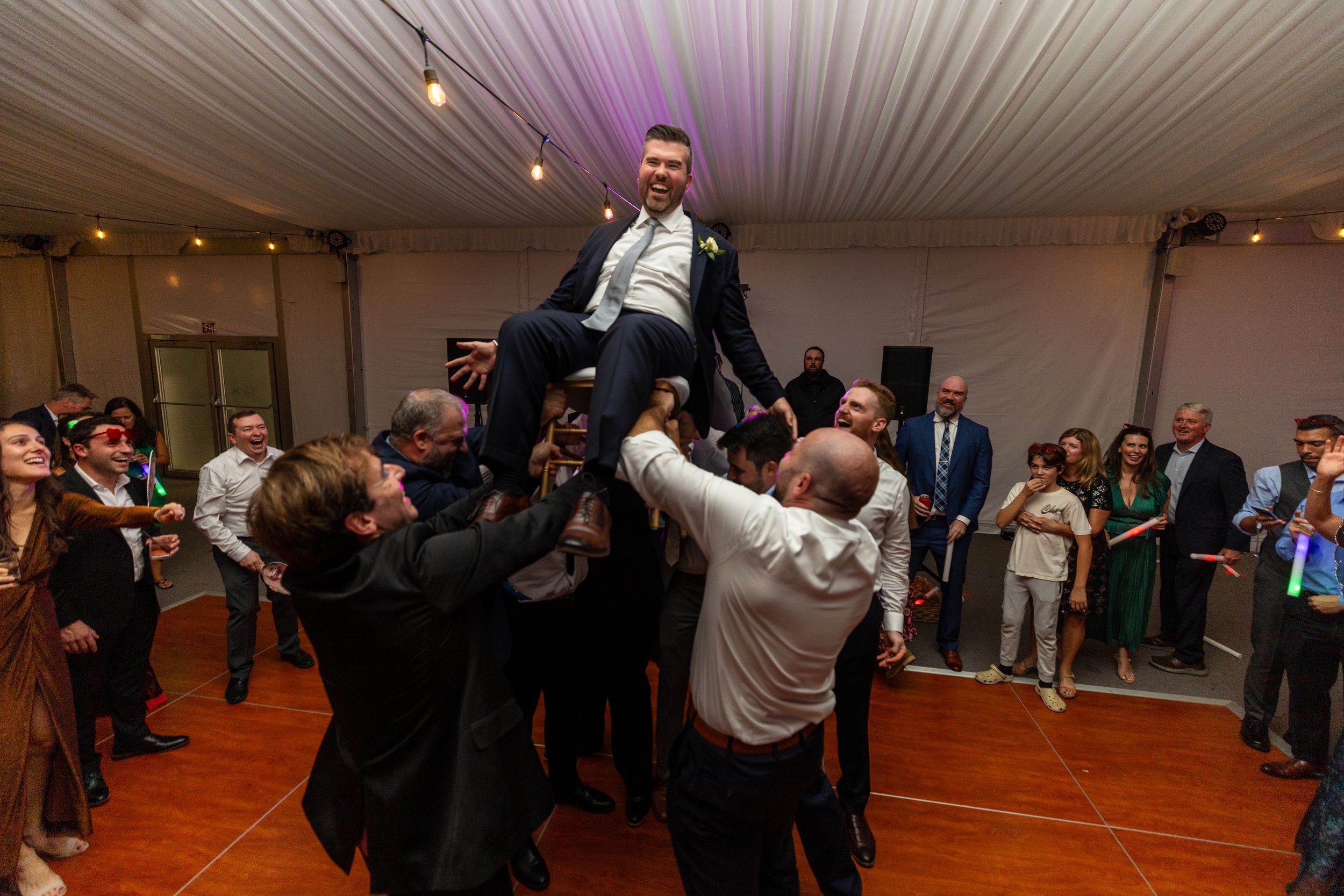 Wedding guests dance and celebrate on a wooden dance floor at an indoor reception venue.