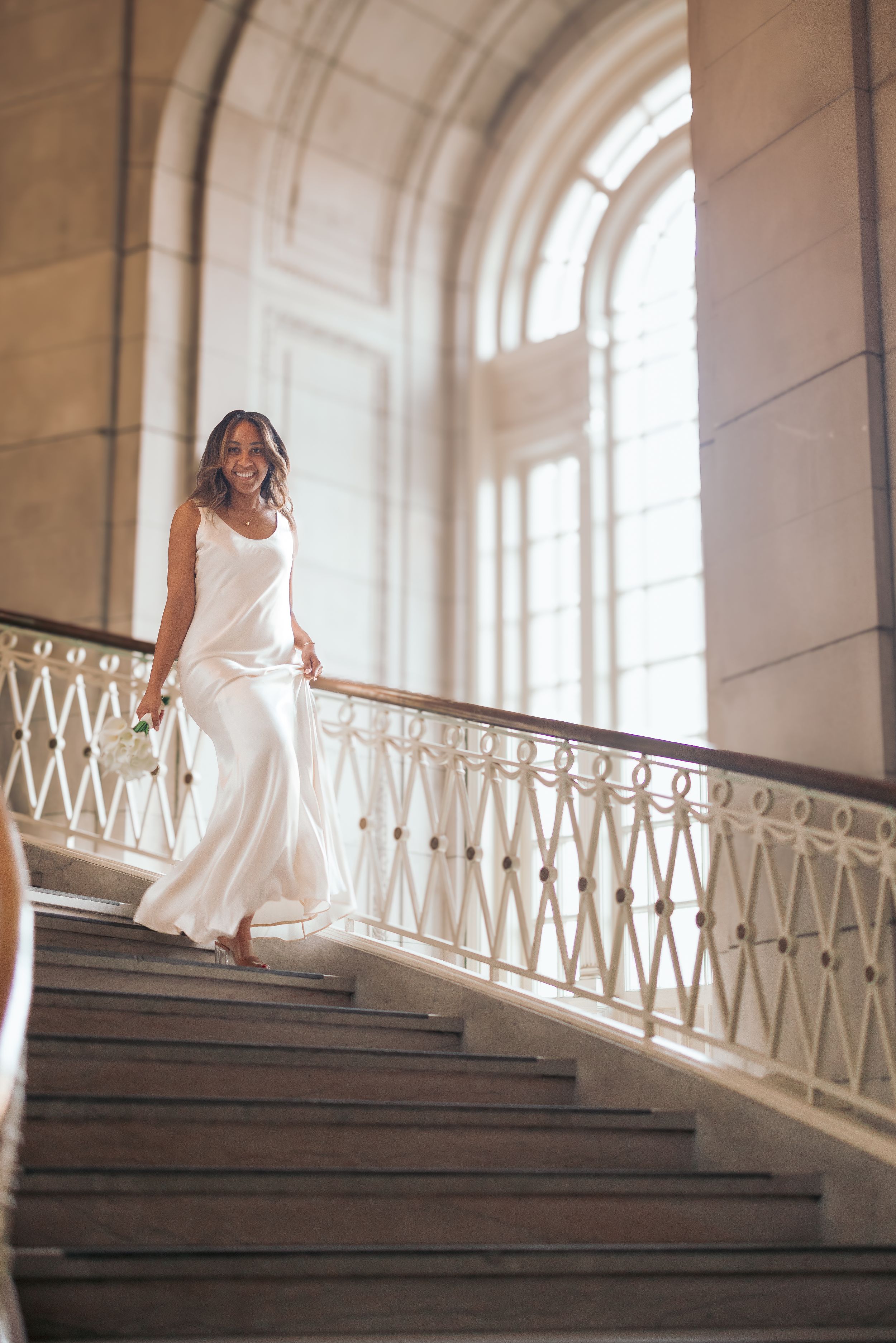 A bride in a white dress descends an elegant marble staircase in a sunlit classical building with arched windows.