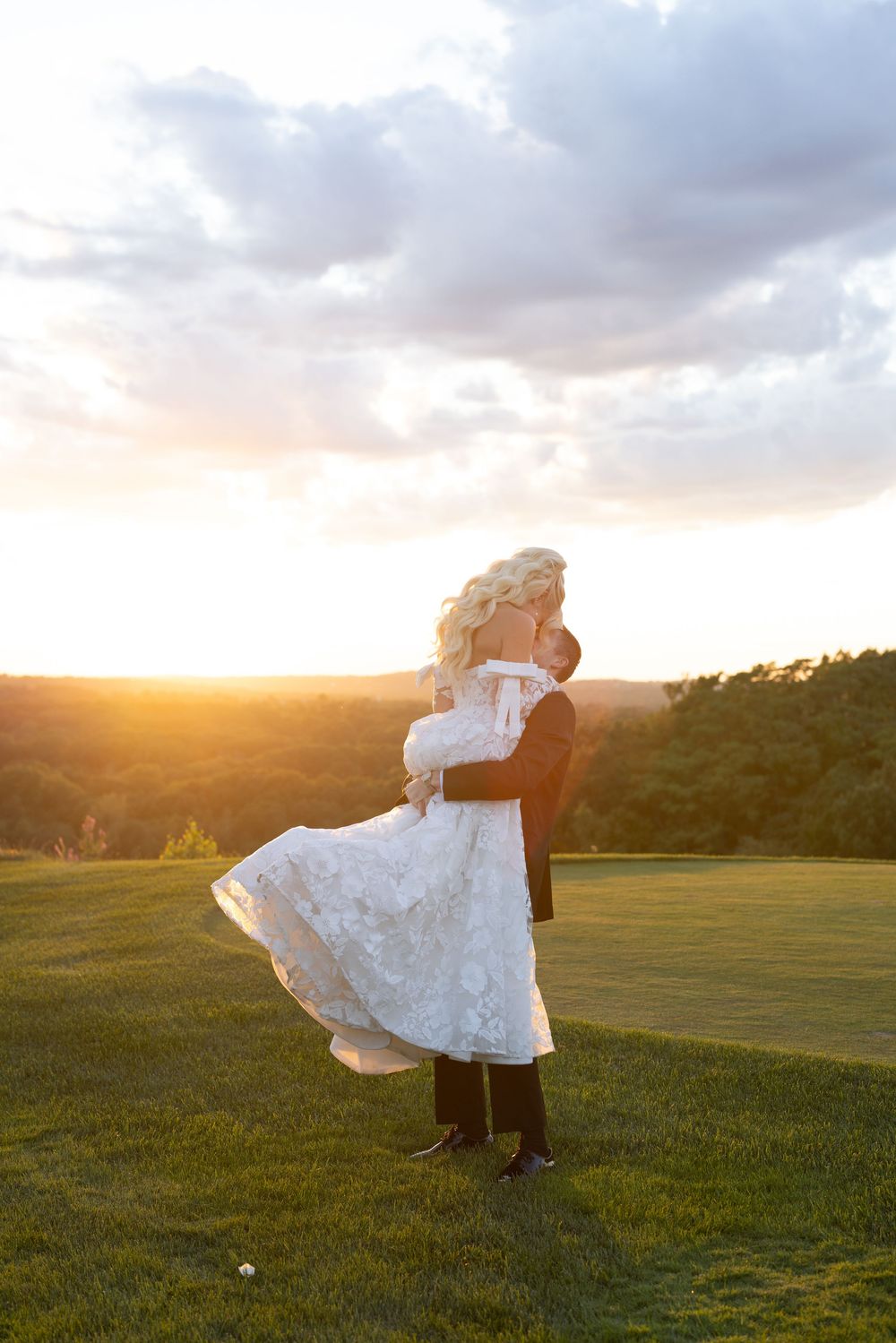 Groom lifting bride in white lace dress at golden sunset on lush green hillside.