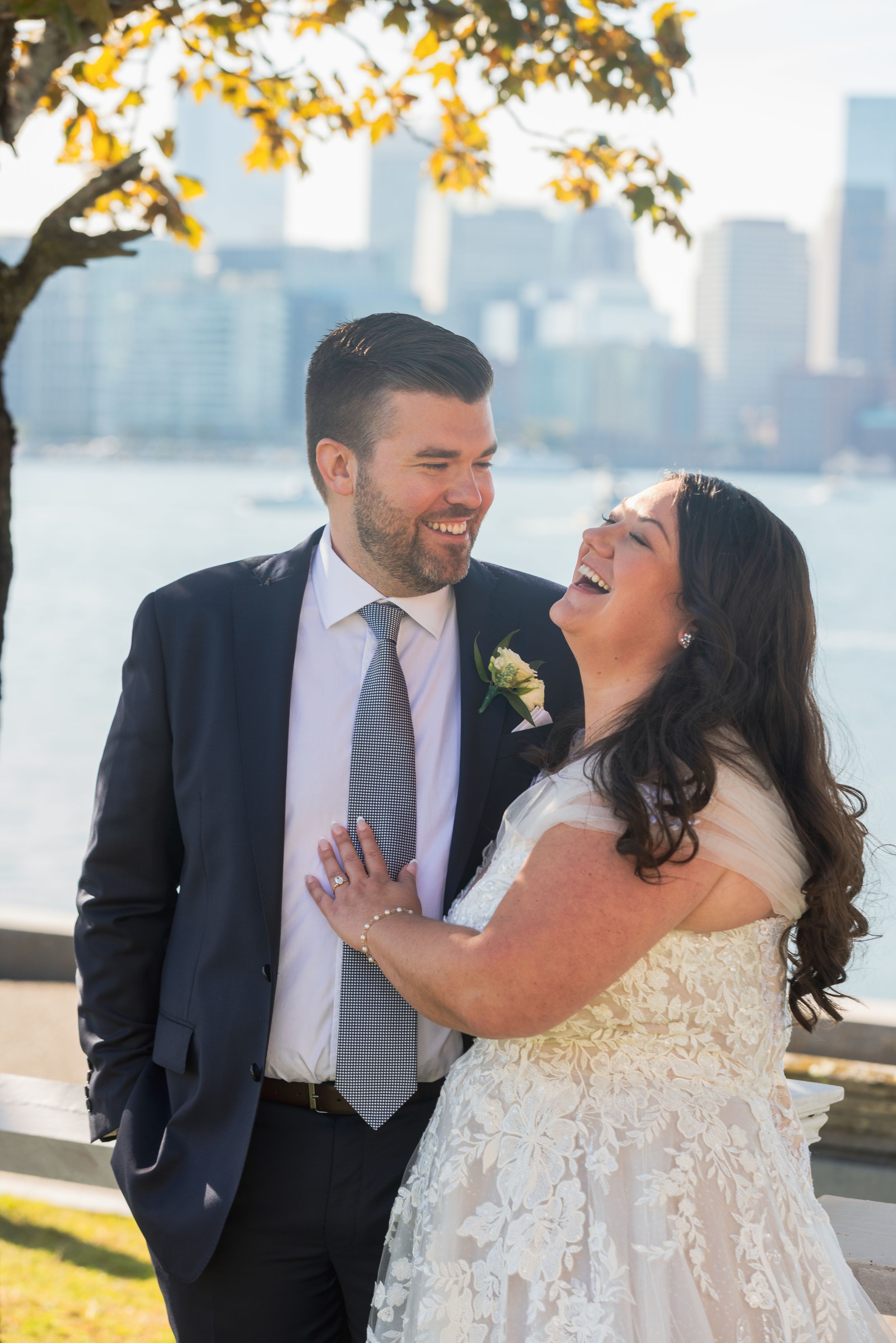 A joyful wedding couple shares a candid laughing moment by the waterfront with a city skyline in the background.