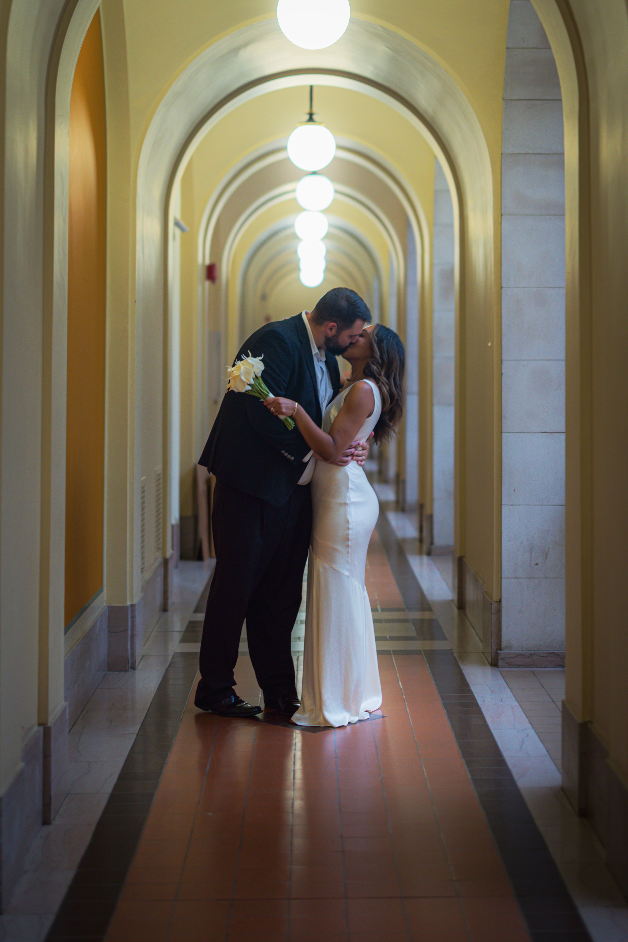 A romantic couple in formal attire shares intimate moments in an elegant arched hallway with glowing pendant lights.
