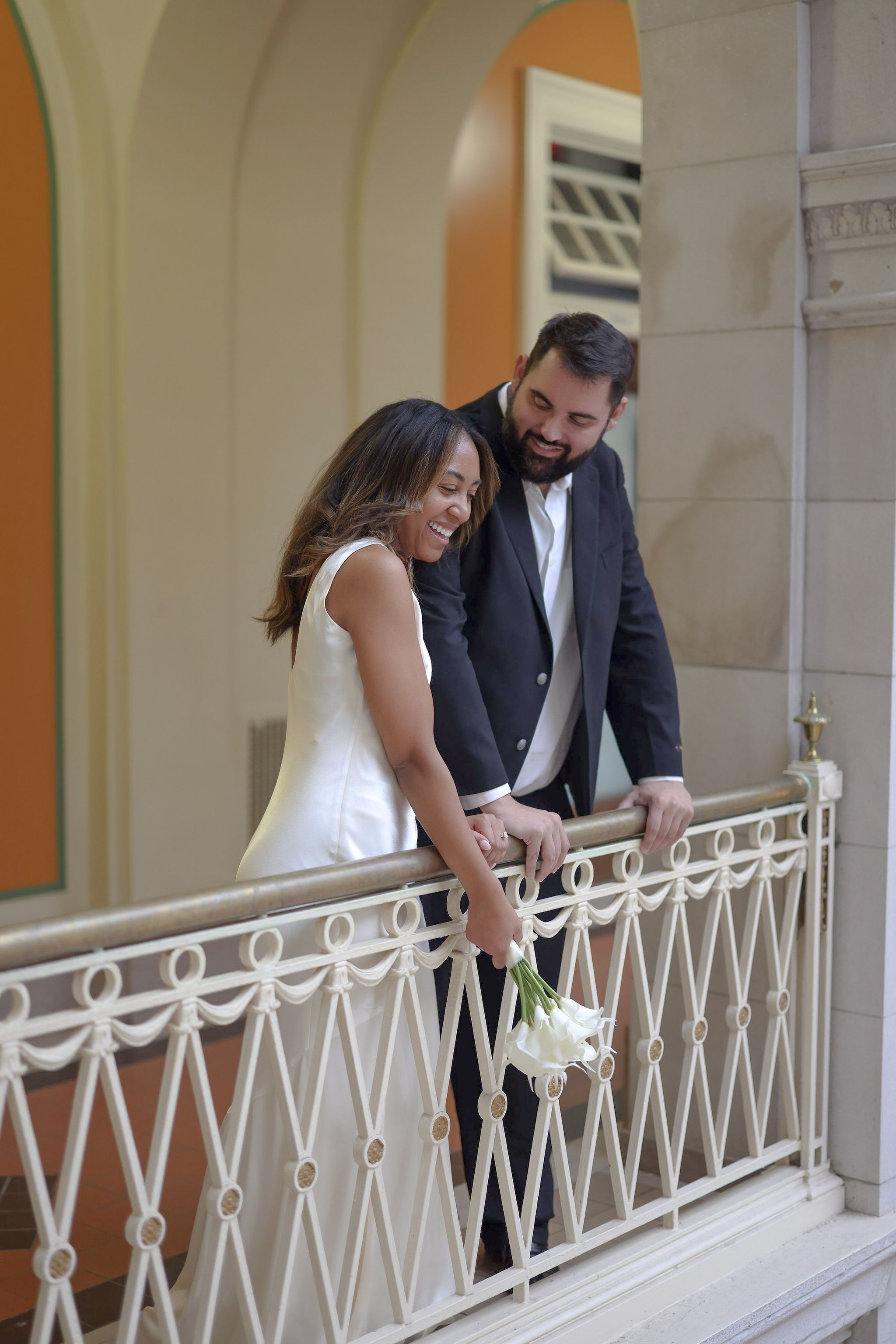 Couple in wedding attire sharing a joyful moment on an elegant white balcony with ornate railing.