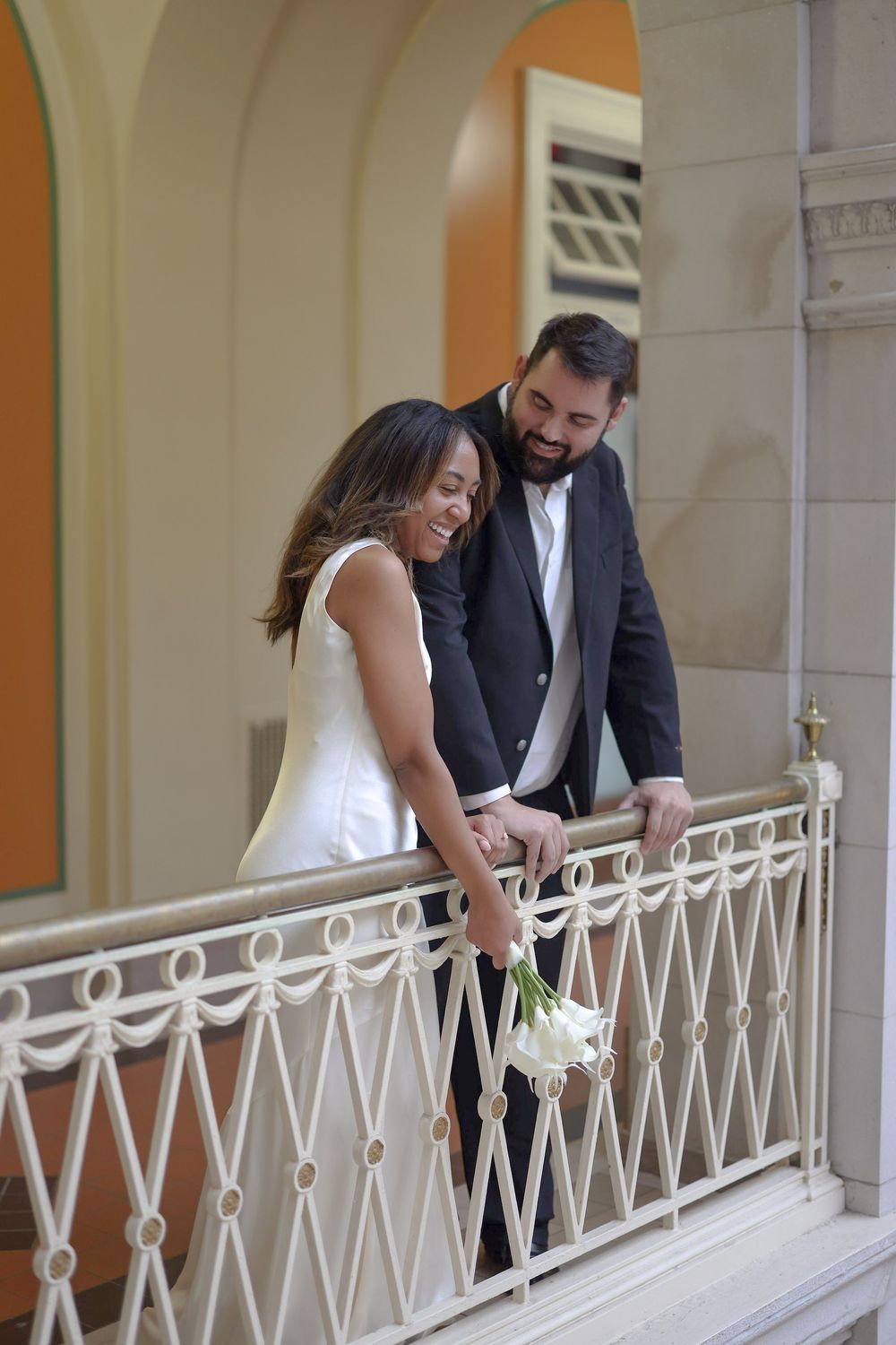 Couple in wedding attire sharing a joyful moment on an elegant white balcony with ornate railing.