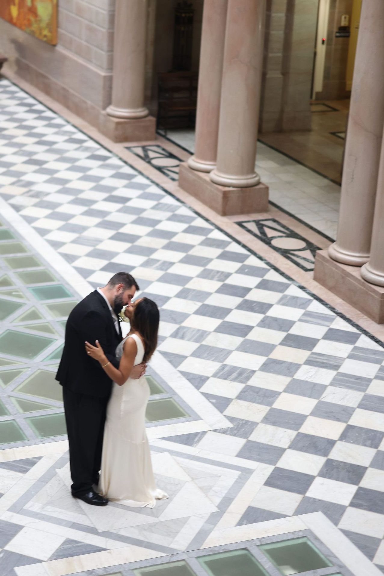 A couple shares a romantic kiss on a checkered marble floor with elegant columns in the background.