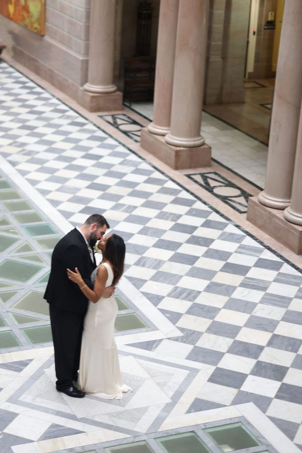 A couple shares a romantic kiss on a checkered marble floor with elegant columns in the background.