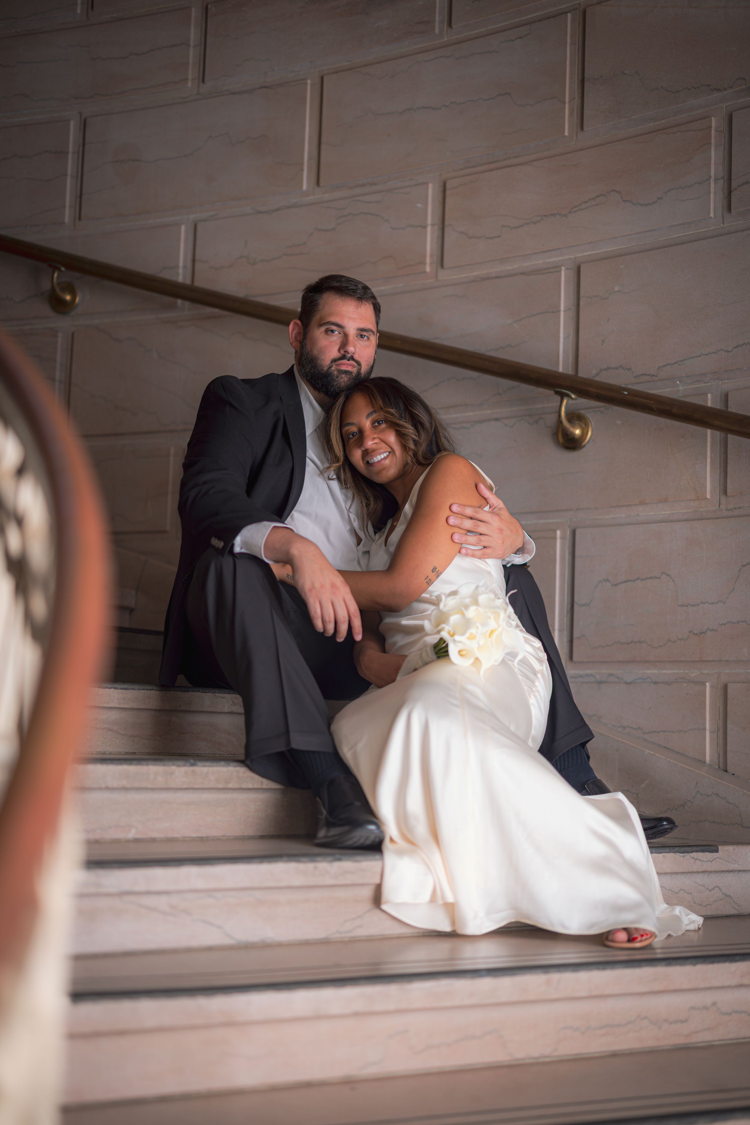 A couple in wedding attire shares an intimate moment while lying on wooden stairs in a rustic setting.