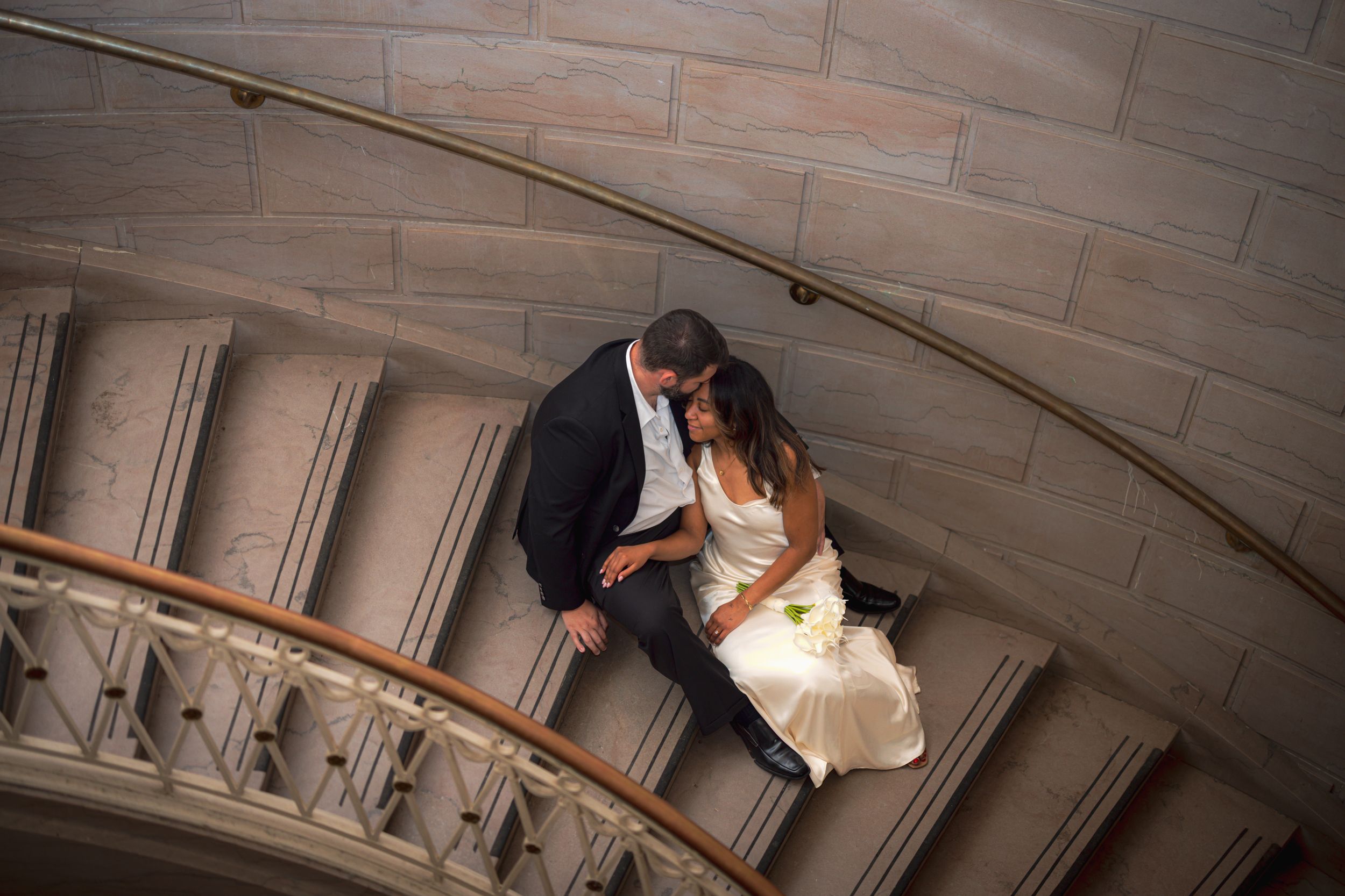 A couple in wedding attire embraces on a grand marble staircase in an elegant architectural setting.