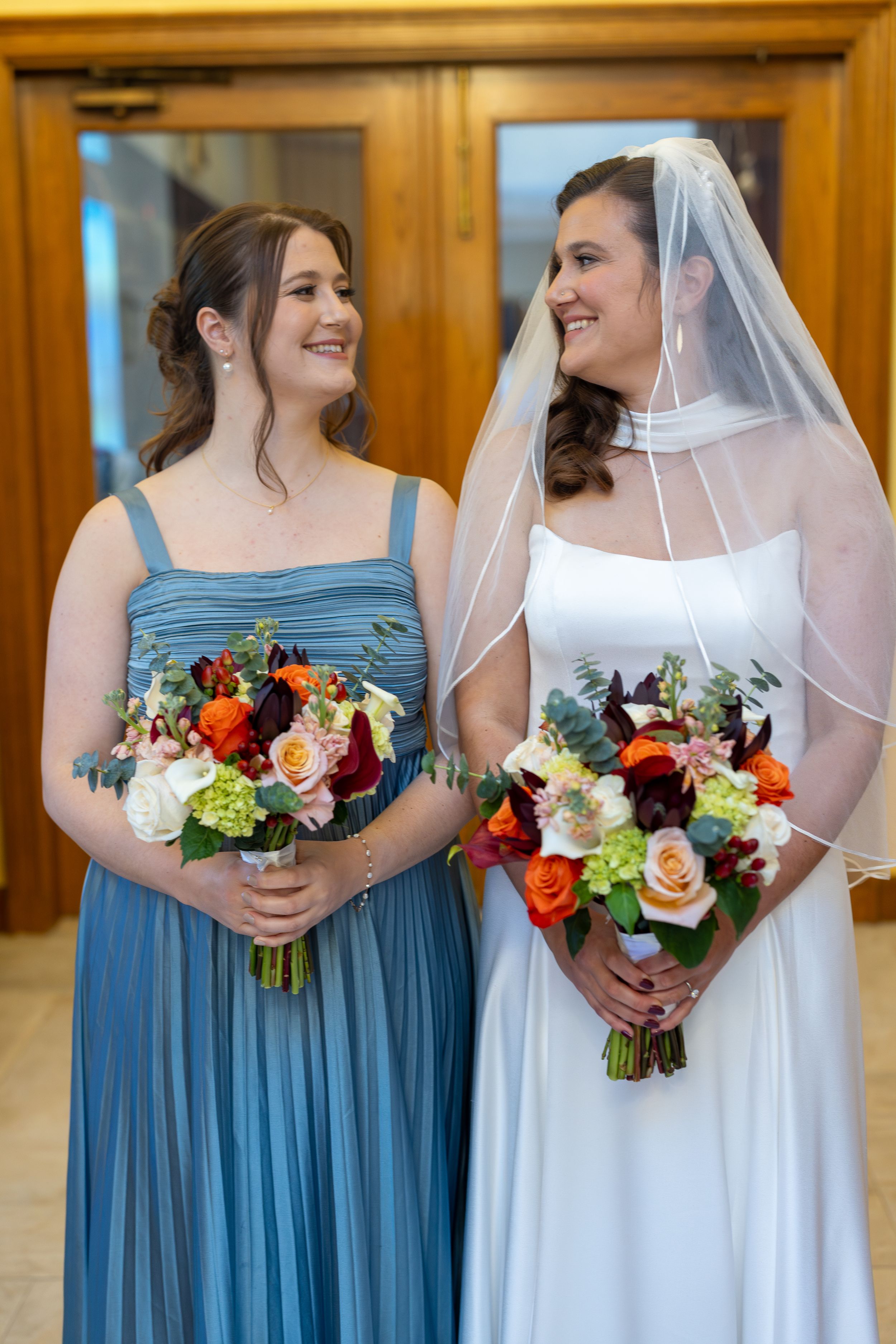 Bridesmaids in dusty blue dresses and bride in white hold vibrant red and peach floral bouquets at wedding ceremony.
