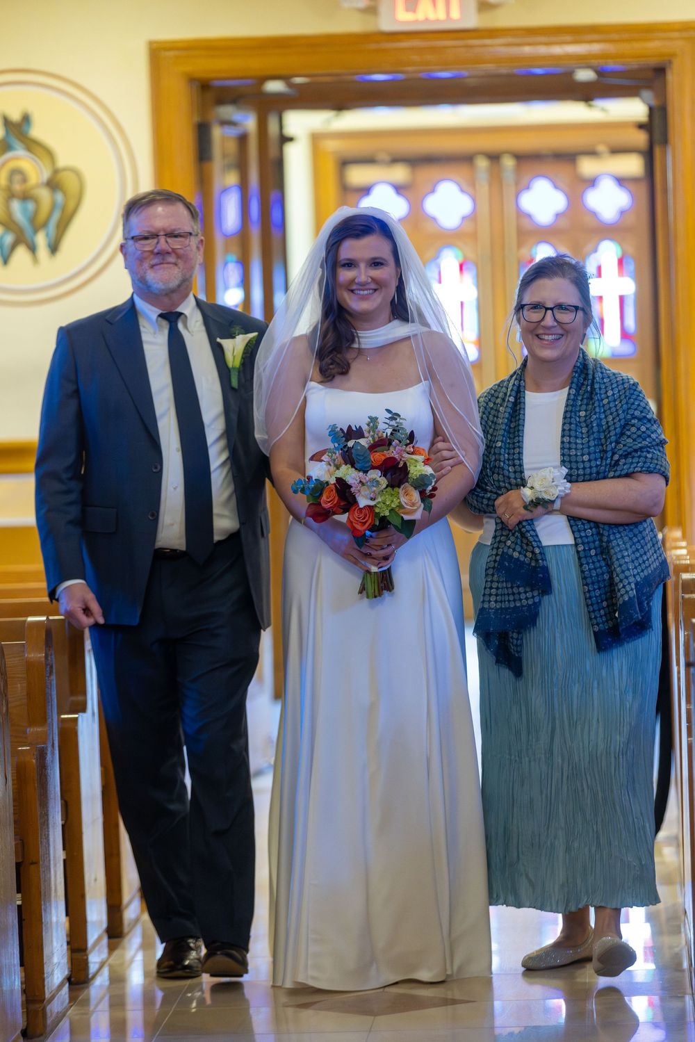 Three people stand together in a church hallway with ornate decor, with the person in the middle wearing a white wedding gown.