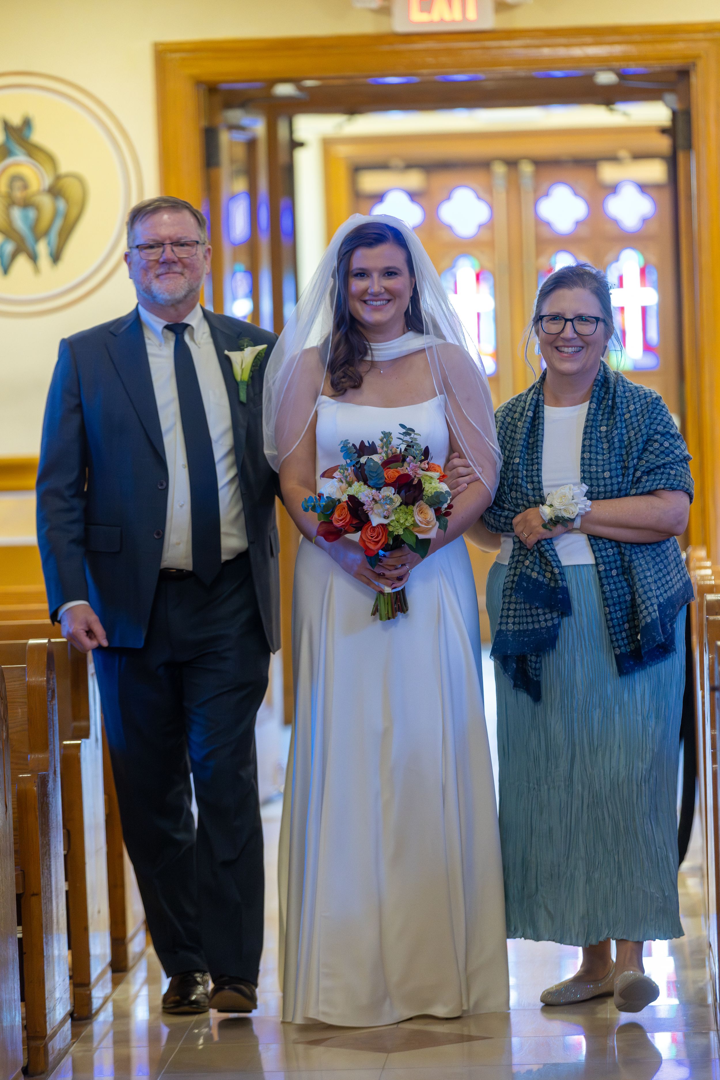Three people stand together in a church hallway with ornate decor, with the person in the middle wearing a white wedding gown.