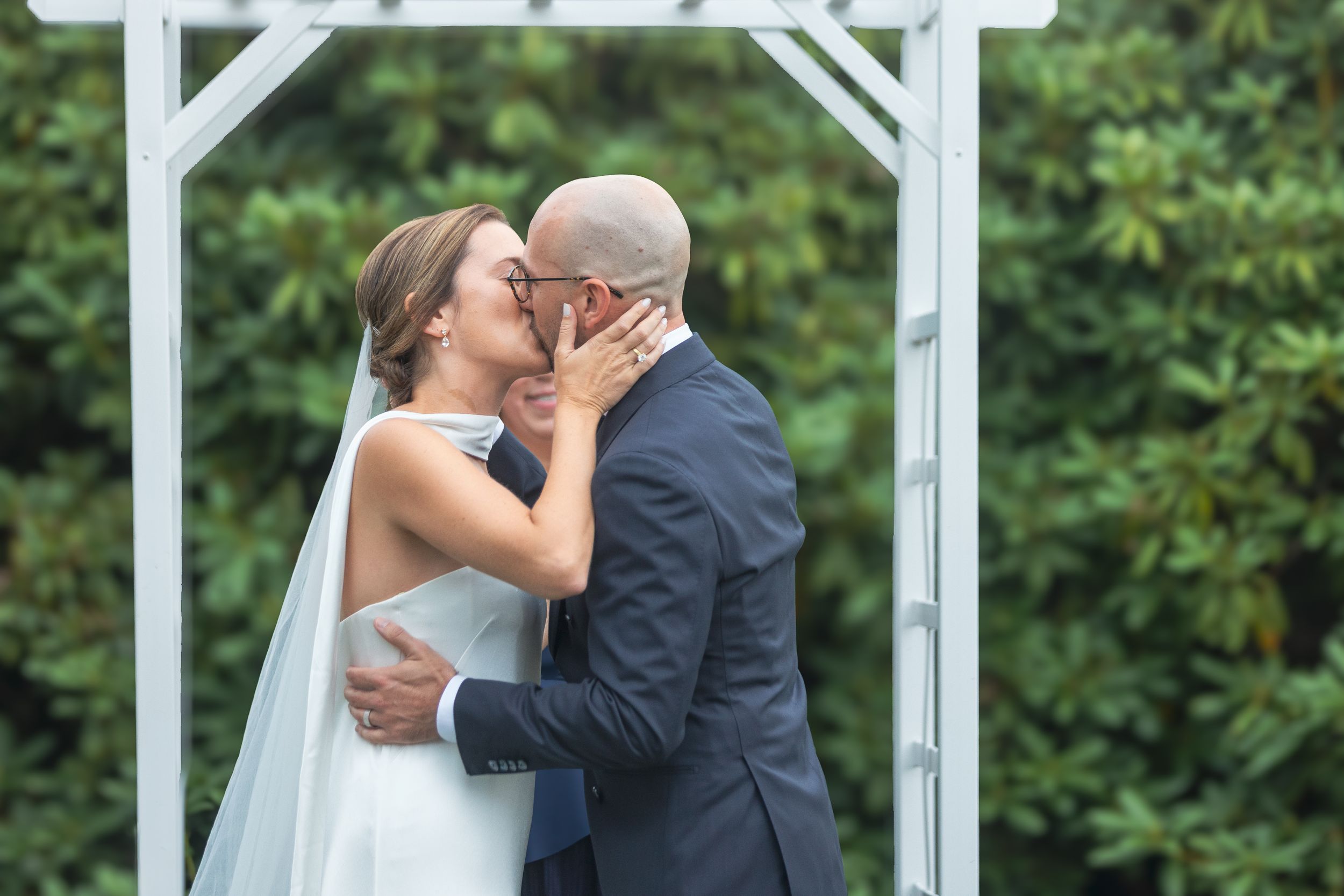 A romantic wedding couple shares a kiss under a white gazebo surrounded by lush greenery.