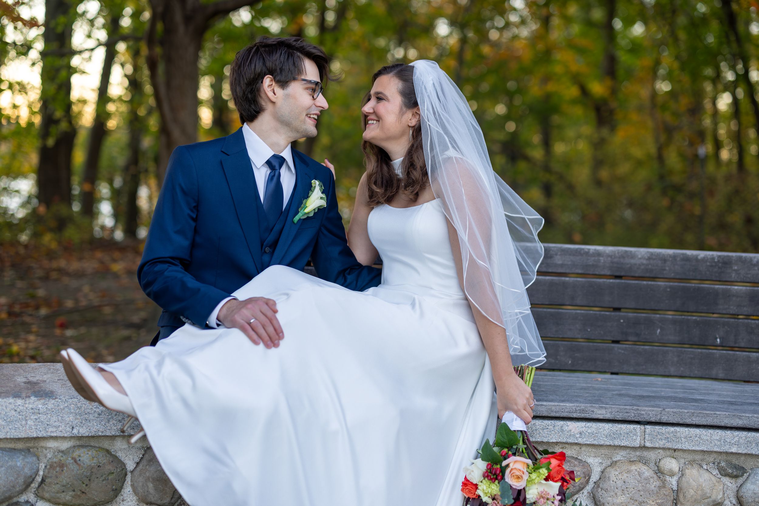 Newlyweds share romantic moments in an autumn forest setting with fall foliage and stone walls.