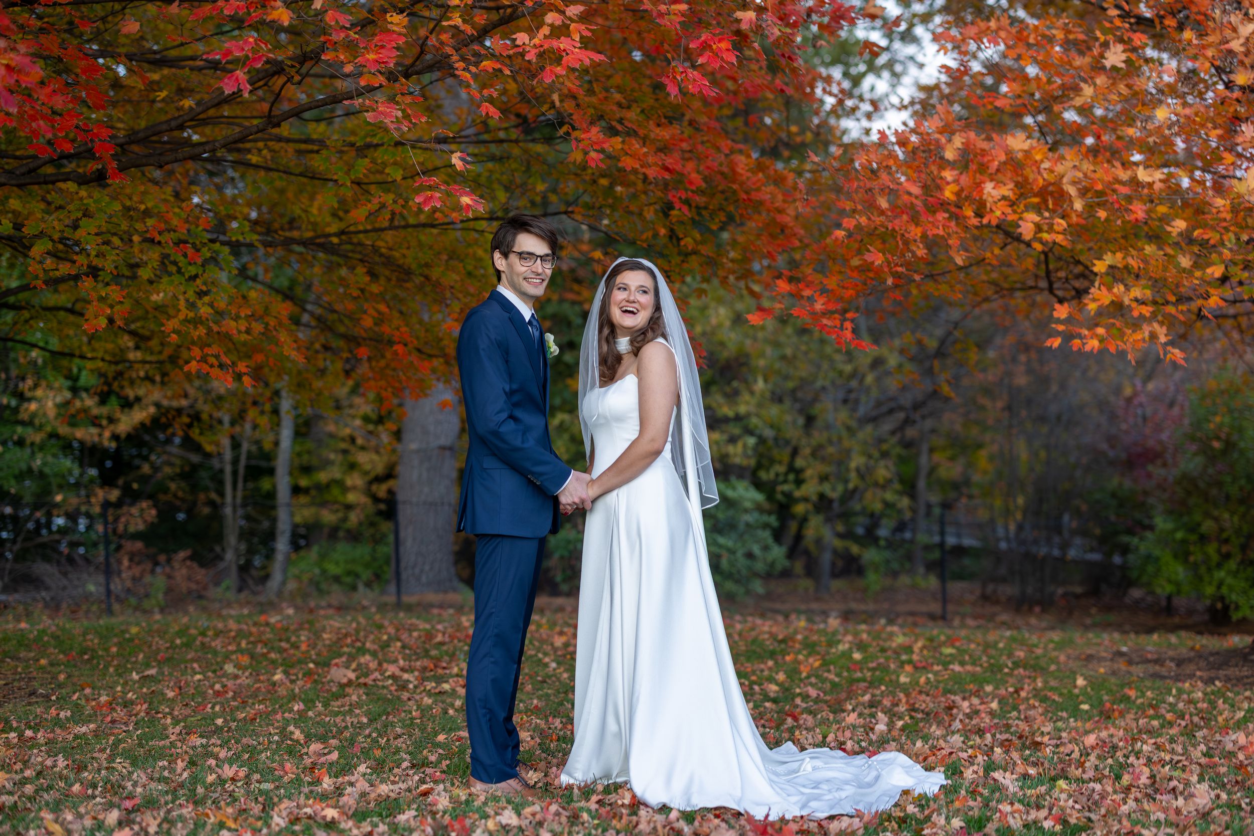 A couple in wedding attire stands together beneath vibrant autumn foliage in a forest setting.