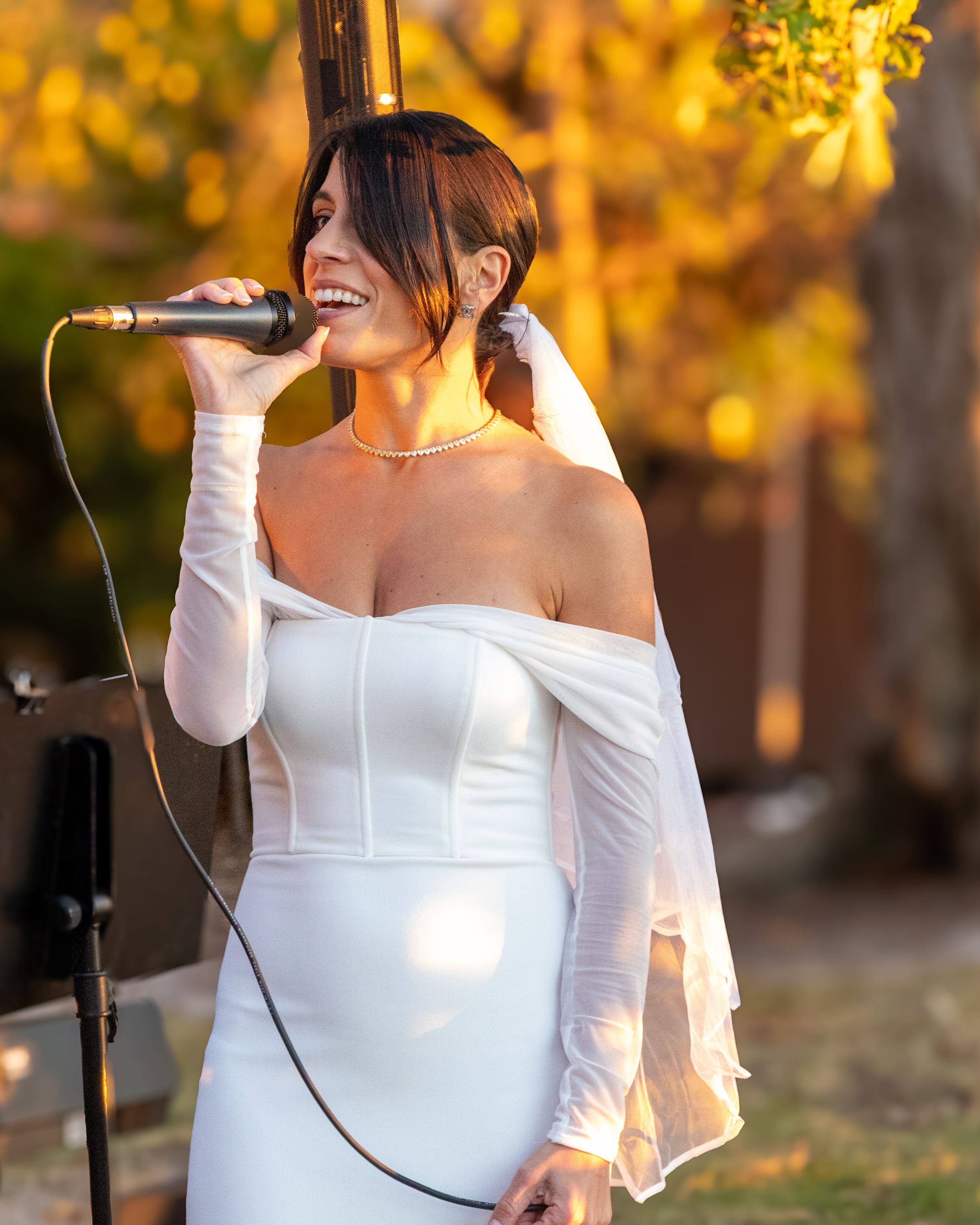 Bride in white off-shoulder dress poses in autumn setting with golden foliage in background.