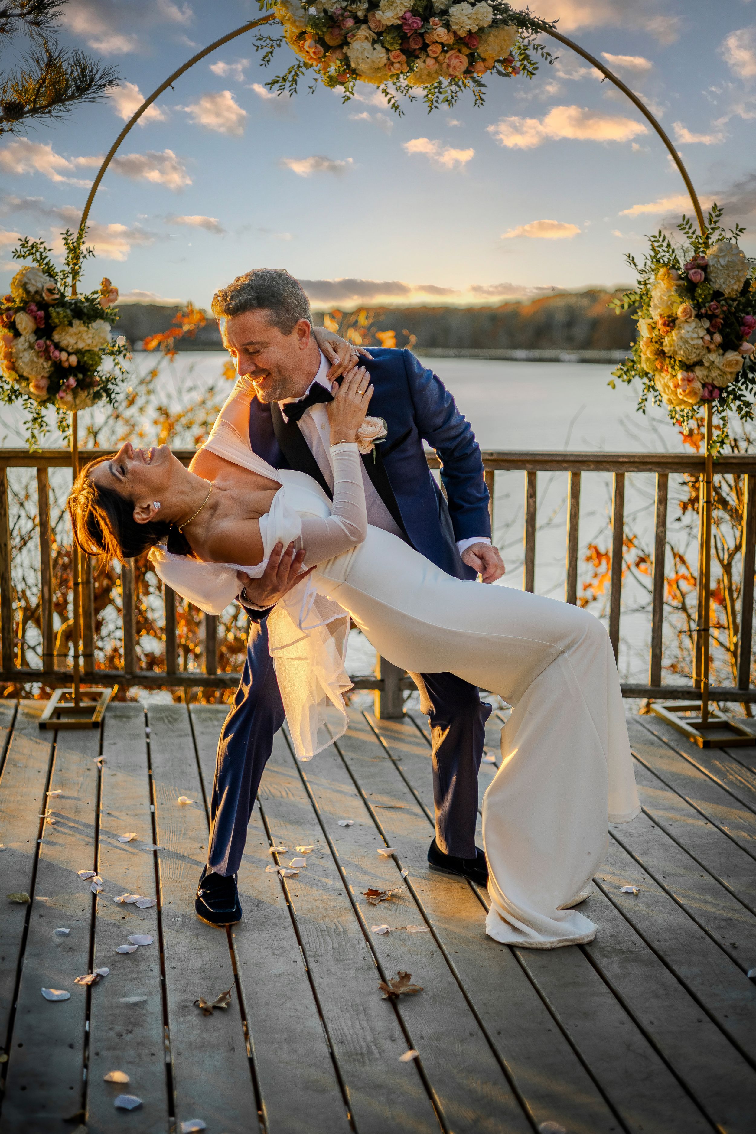 Romantic wedding couple dips under floral arch on lakeside deck during golden sunset.